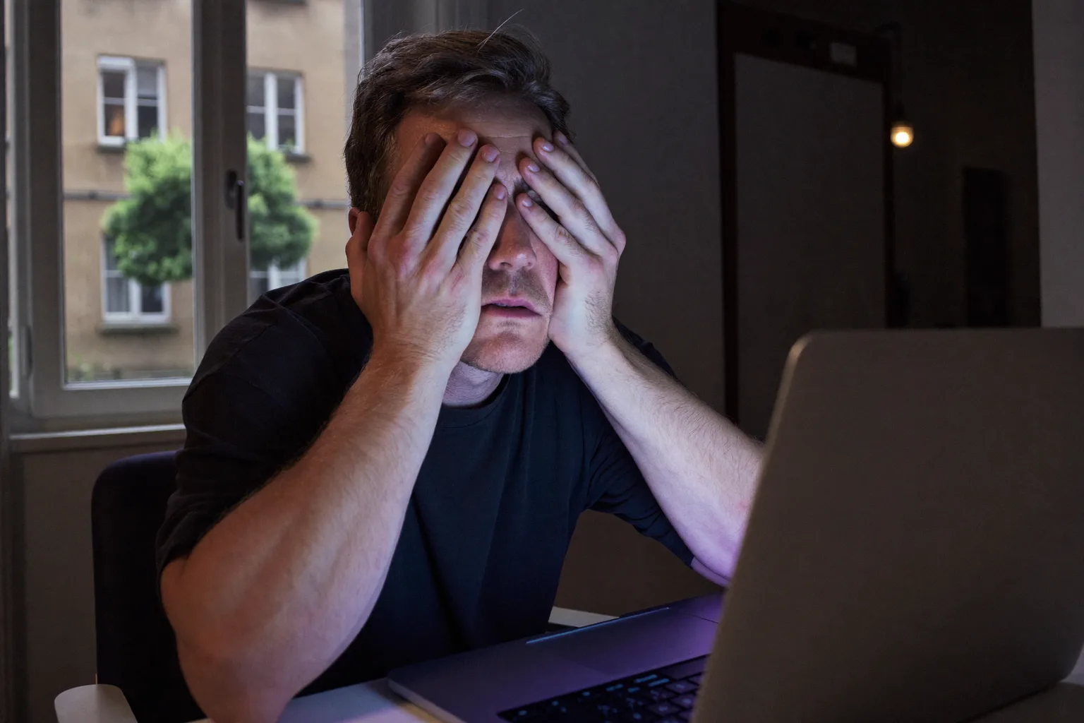 Egy férfi az asztalánál, félhomályos szobában, tenyerével a csukott szemét nyomja, hogy enyhítse a szemfáradtságot, arcát a laptop képernyőjének fénye világítja meg