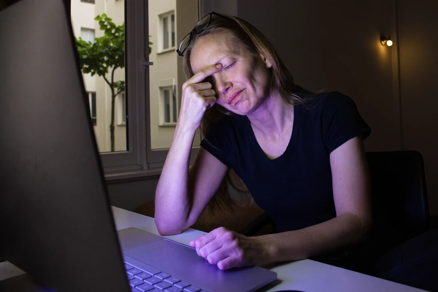 Egy nő az íróasztalánál egy sötét szobában, csukott szemmel, szemüvegét a homlokára tolva, az orrnyergét masszírozza a monitor mögött, arcát hidegen kékes fény világítja meg a képernyőről