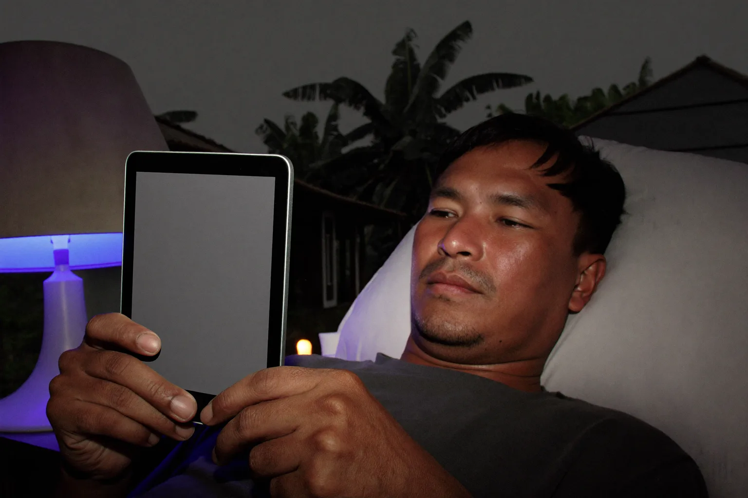 Seorang pria membaca e-reader di tempat tidur larut malam, cahaya halamannya redup dan nyaris tak menyentuh wajahnya, sementara di meja nakas sebelahnya sebuah ponsel yang tergeletak menghadap ke atas membanjiri kap lampu kedua dengan cahaya biru yang keras dan dingin