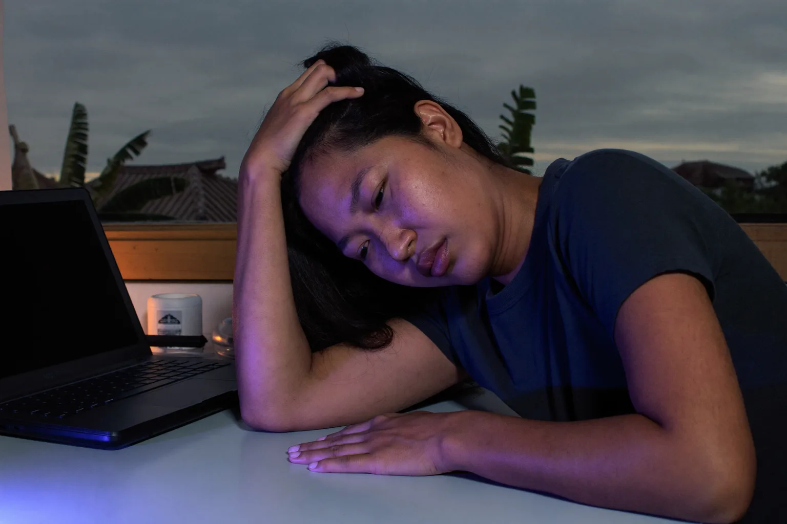 Seseorang di meja kerja rumah pada larut malam, kepala miring bersandar di satu tangan dengan jari-jari di rambutnya, mata menunduk dan tidak fokus, lengan bawah yang lain diam di atas meja yang kosong