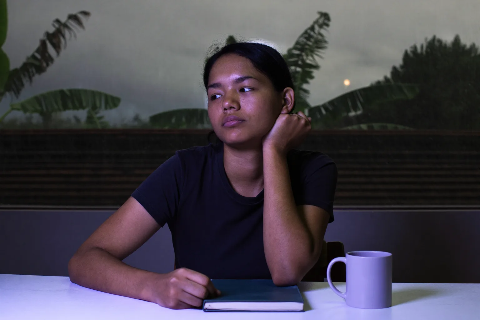 Seorang pelajar di meja belajar larut malam, dagu bertumpu di satu telapak tangan yang terbuka, wajah tersorot cahaya layar dari samping, pandangan menerawang menjauh dari tugas dan keluar bingkai
