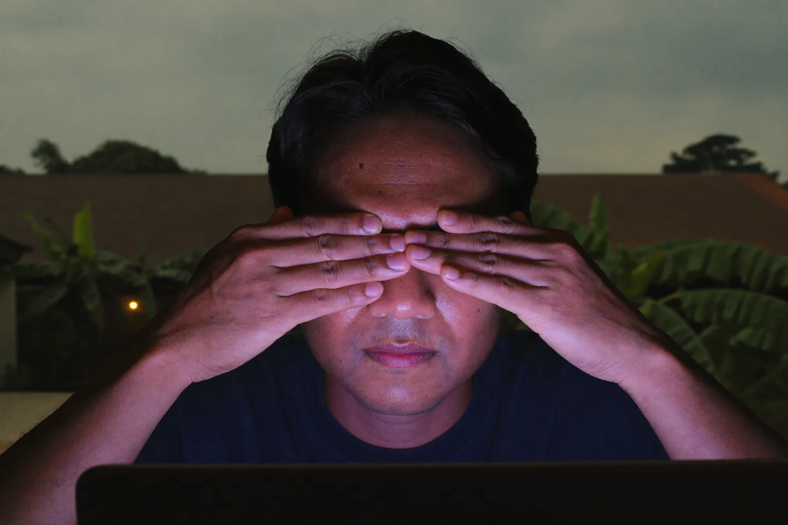 Seorang pria di meja kerja dalam ruangan redup menutup matanya yang terpejam dengan telapak tangan untuk meredakan mata lelah, wajahnya diterangi cahaya layar laptop