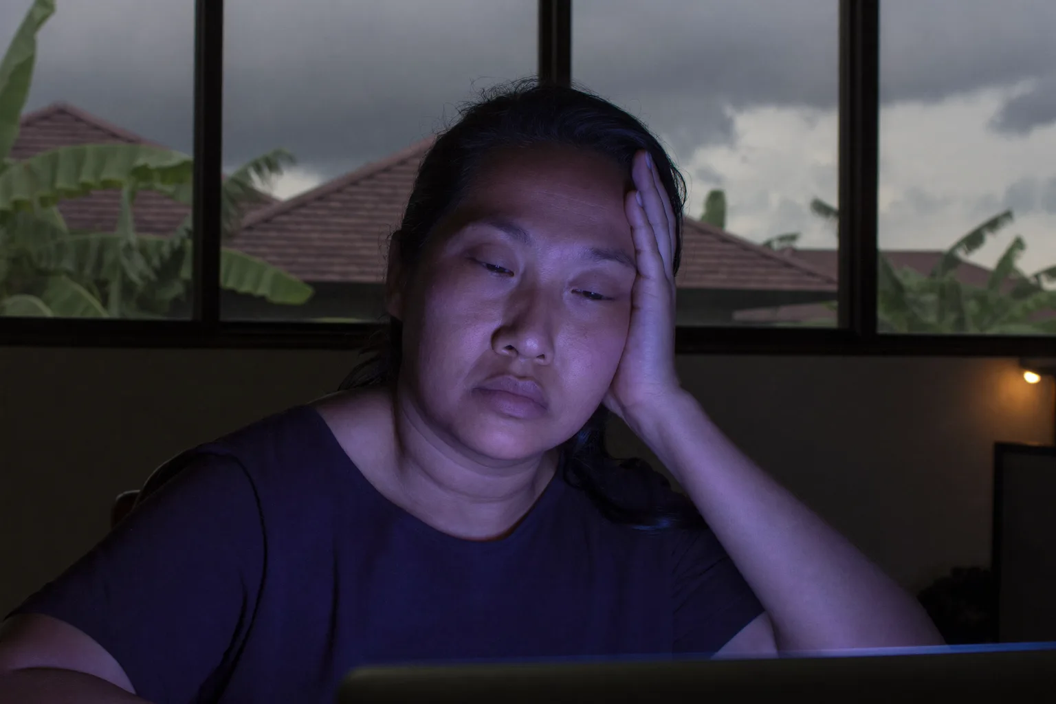 Seorang wanita lelah di depan laptop menjelang malam, bersandar dengan tangan menopang kepala, matanya berat dan kering dengan lingkaran hitam di ruangan yang temaram