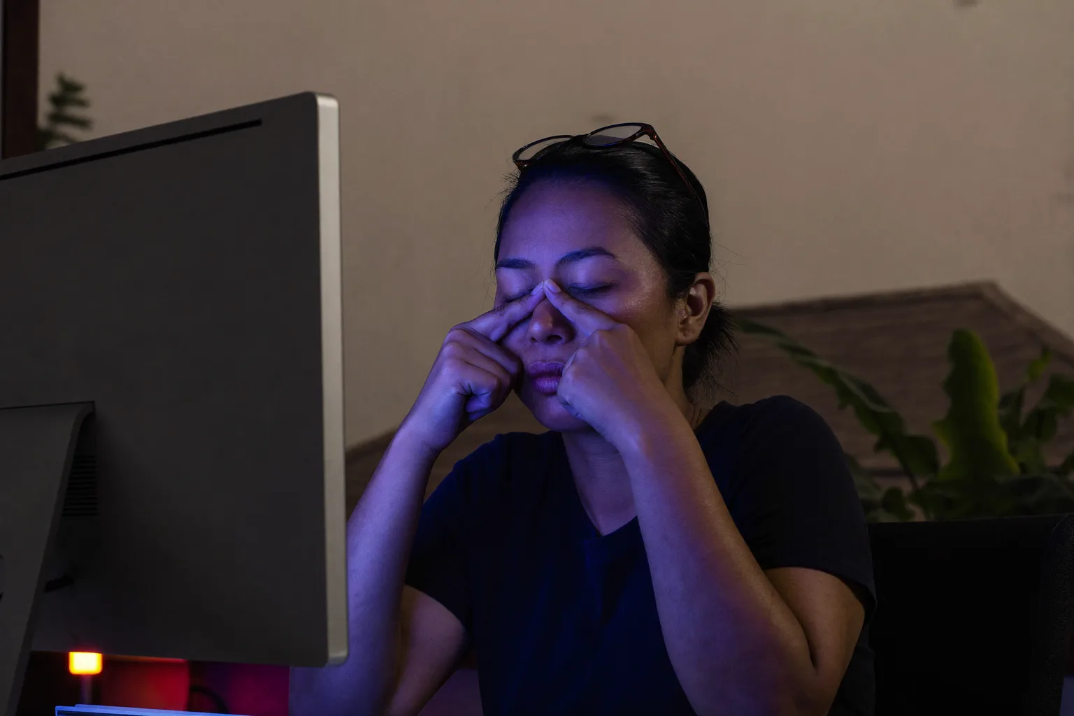 Seorang wanita di meja kerja dalam ruangan gelap, matanya terpejam dan kacamatanya didorong ke atas dahi, memijat pangkal hidungnya di belakang monitor, wajahnya diterangi cahaya biru dingin dari layar