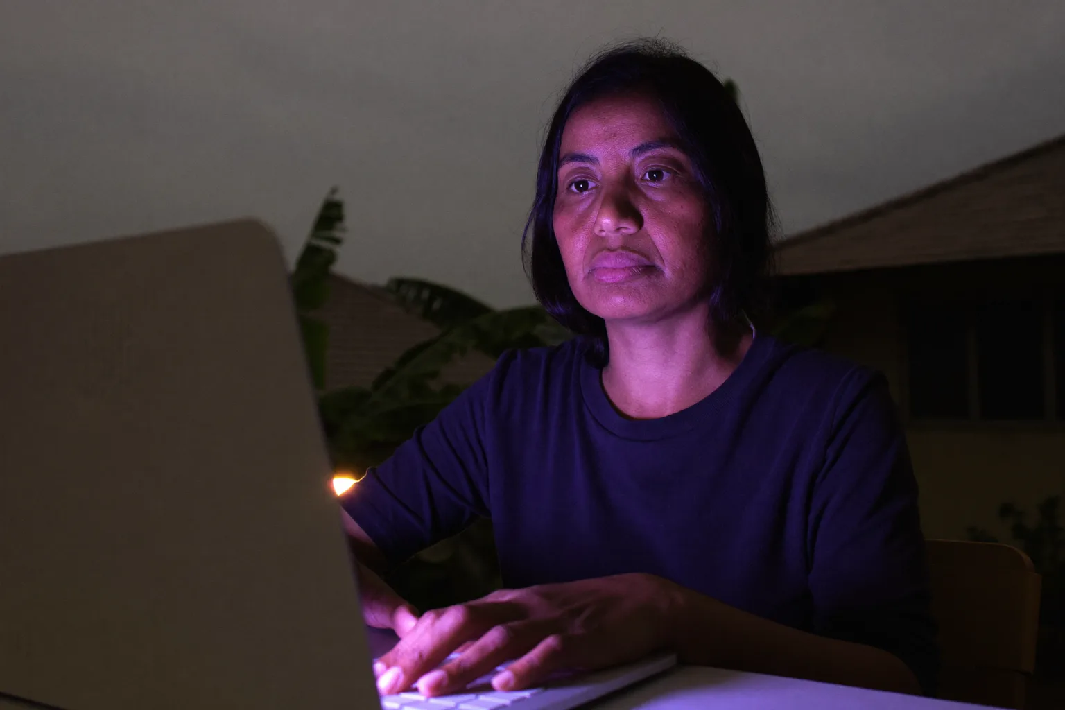 Seseorang duduk di meja kerja rumah larut malam, menatap kosong melewati layar laptop dengan tangan masih di keyboard, menunjukkan kelelahan datar akibat burnout kerja