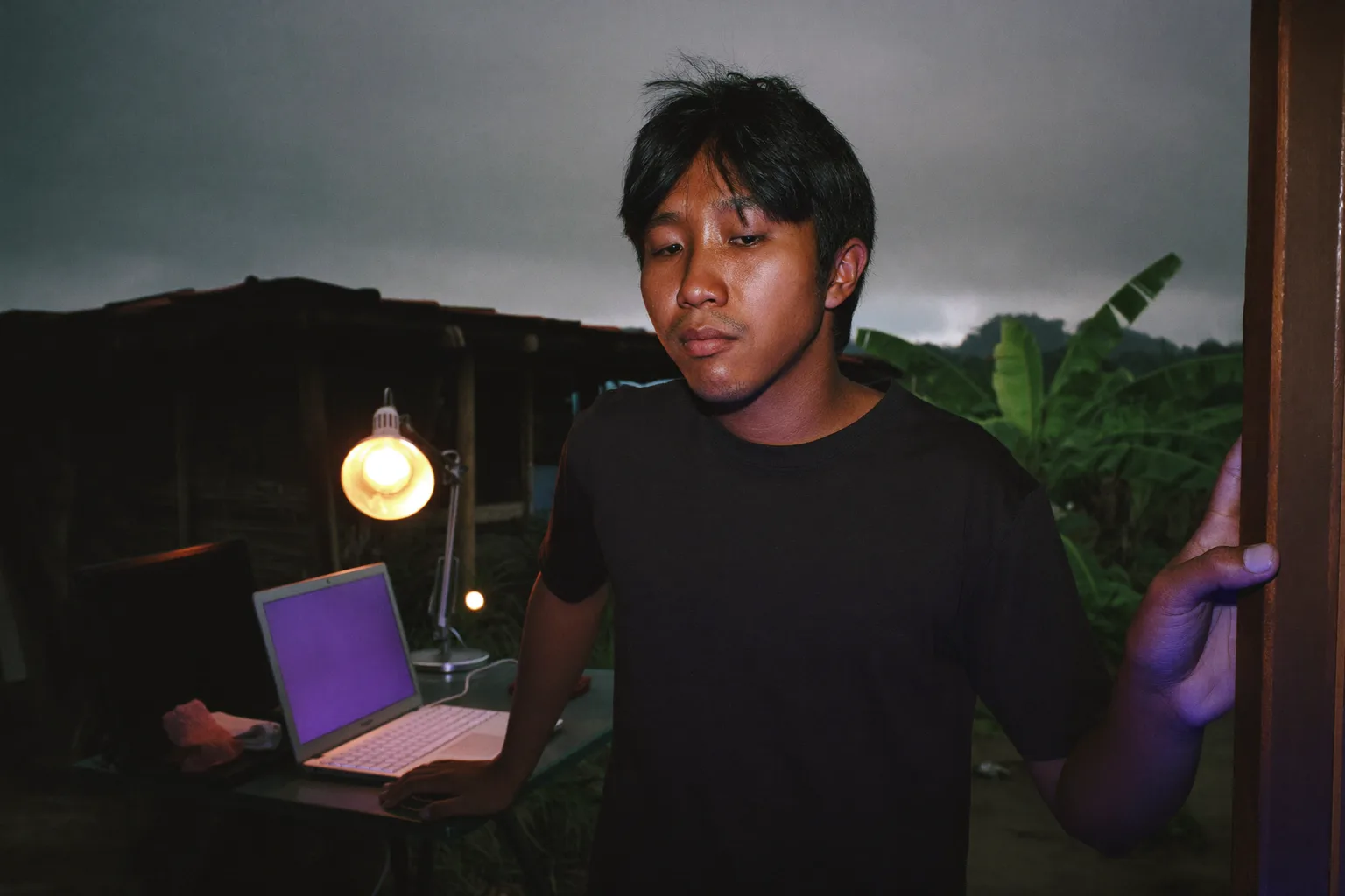 Laptop yang tertutup di meja kerja rumah pada malam hari dengan lampu ruangan menyala dan orangnya berjalan menjauh, menggambarkan akhir dari hari kerja jarak jauh