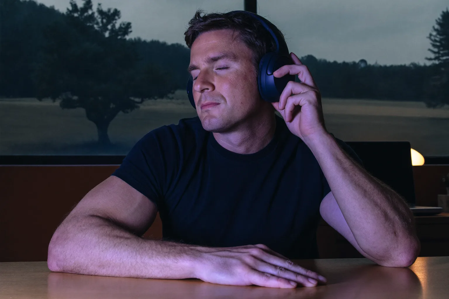 A person at a desk wearing over-ear headphones, eyes closed and one hand resting on the ear cup, deep in a work session with music playing