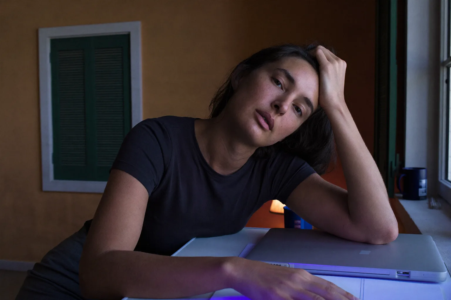 Una persona a una scrivania di casa a tarda sera, con la testa reclinata su una mano e le dita tra i capelli, lo sguardo basso e assente, l’altro avambraccio immobile su una scrivania vuota