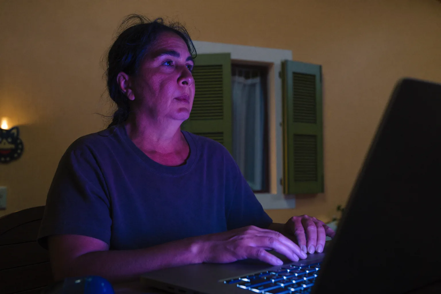 Una persona seduta alla scrivania di casa a tarda sera, con lo sguardo perso oltre lo schermo del laptop e le mani ancora sulla tastiera, che mostra la stanchezza piatta del burnout sul lavoro