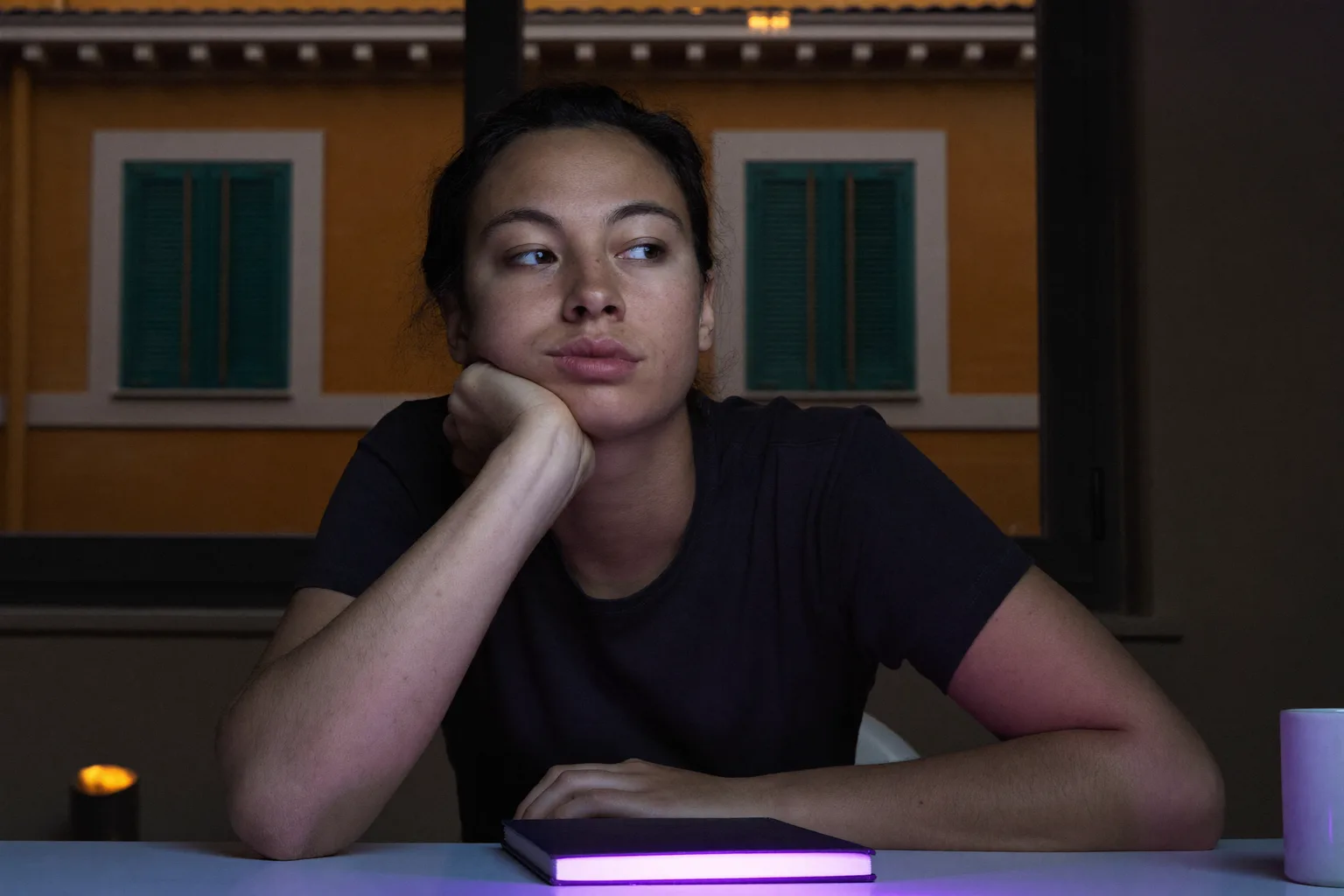 Uno studente alla scrivania a tarda sera, mento appoggiato su una mano aperta, viso illuminato di lato dal bagliore dello schermo, sguardo che si è allontanato dal lavoro e fuori dall’inquadratura