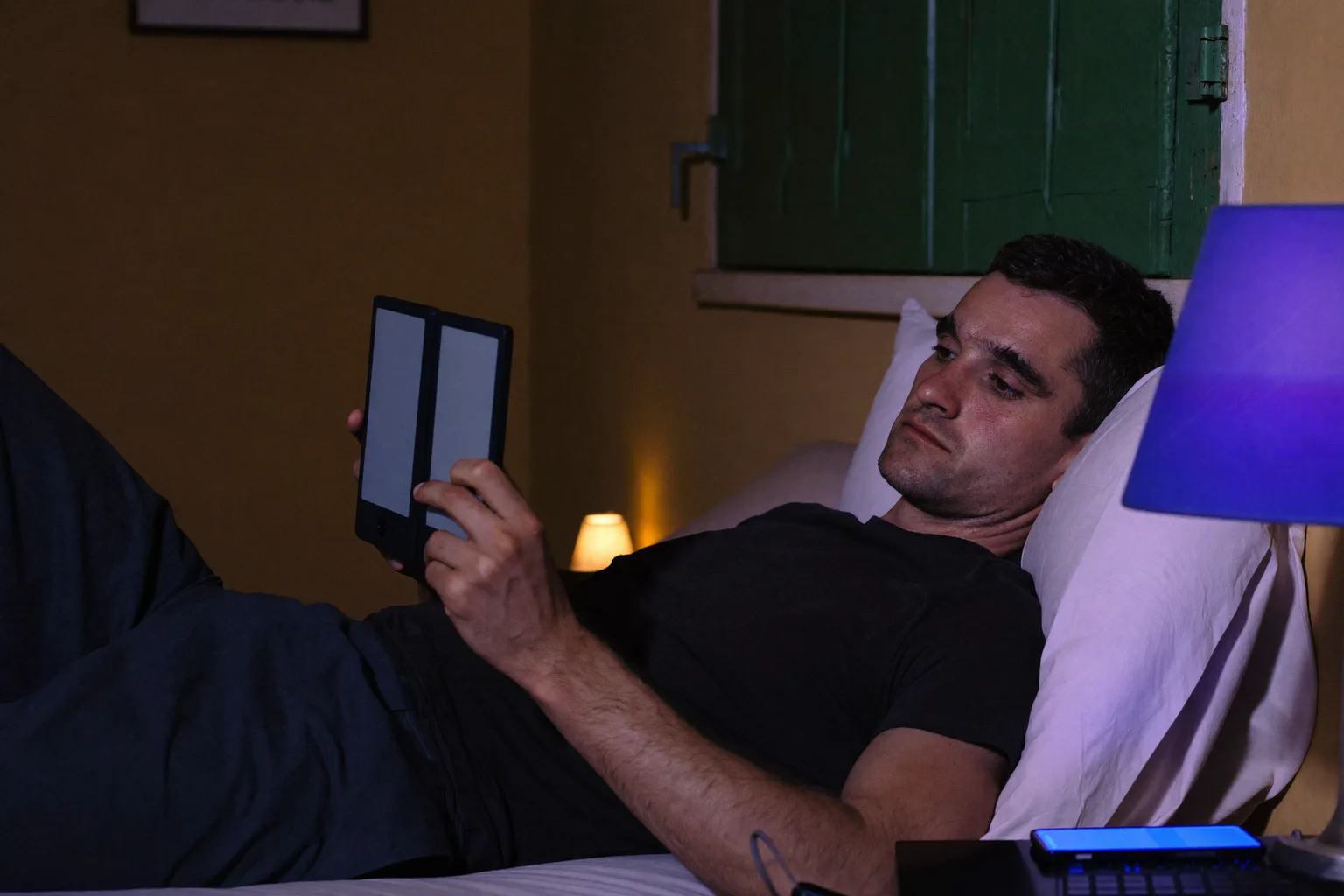 Un uomo che legge da un e-reader a letto, a notte fonda, la pagina un bagliore fioco che gli arriva appena al viso, mentre sul comodino accanto a lui un telefono appoggiato a schermo in su inonda il secondo paralume di una luce blu dura e fredda