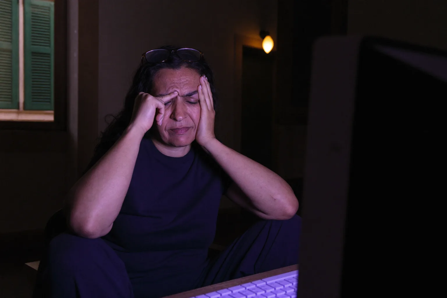 Una donna alla scrivania in una stanza buia, occhi chiusi e occhiali spinti sulla fronte, che si pizzica il ponte del naso dietro al monitor, il viso illuminato da una fredda luce blu proveniente dallo schermo