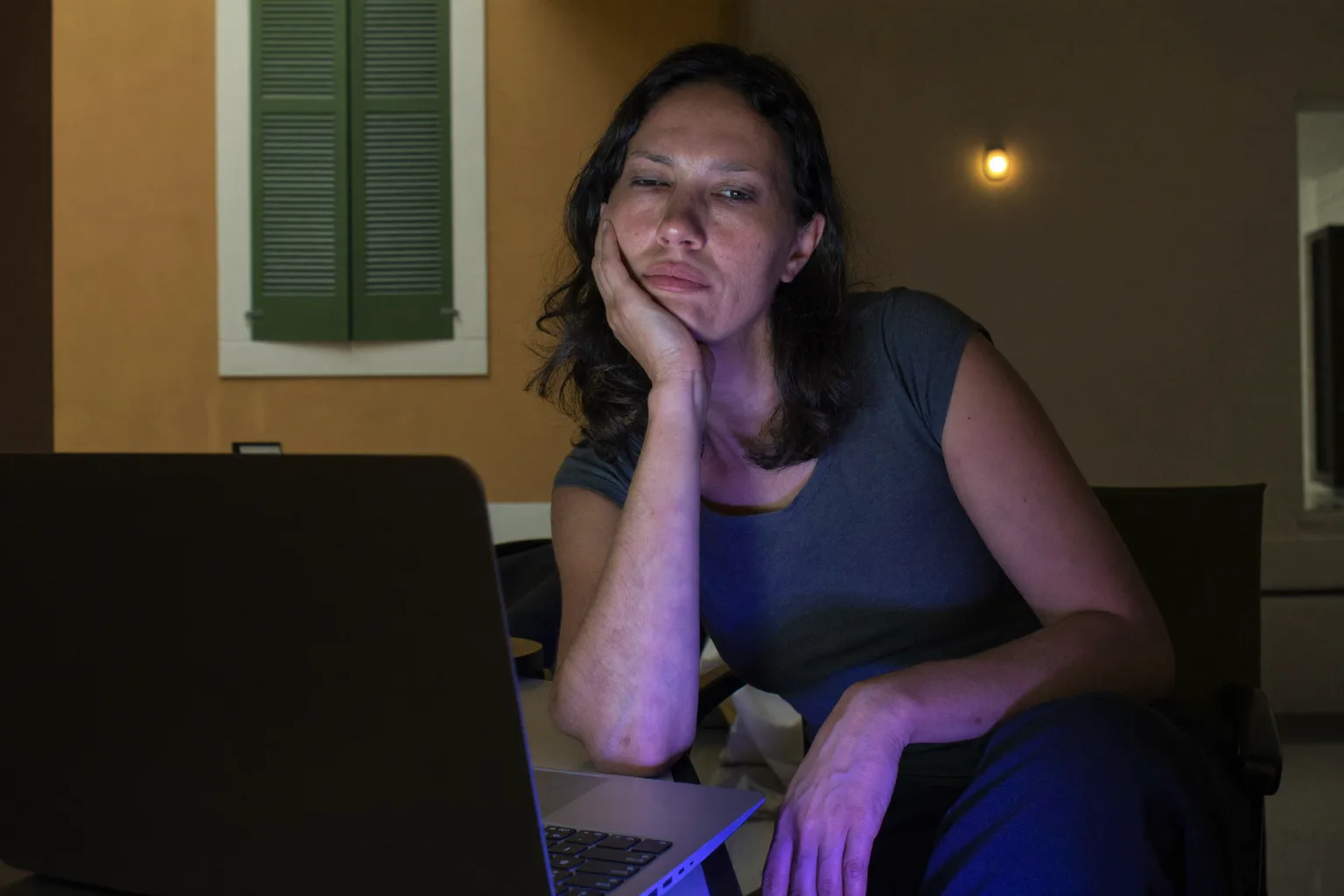 Una donna stanca al computer portatile a fine giornata, con la testa appoggiata su una mano, gli occhi pesanti e secchi e le occhiaie, in una stanza con poca luce