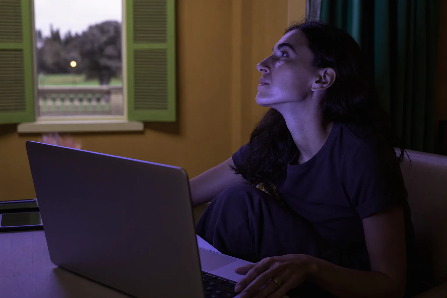 Persona al lavoro al computer che applica la regola del 20-20-20 contro l’affaticamento degli occhi, distogliendo lo sguardo dal laptop verso un albero lontano oltre la finestra