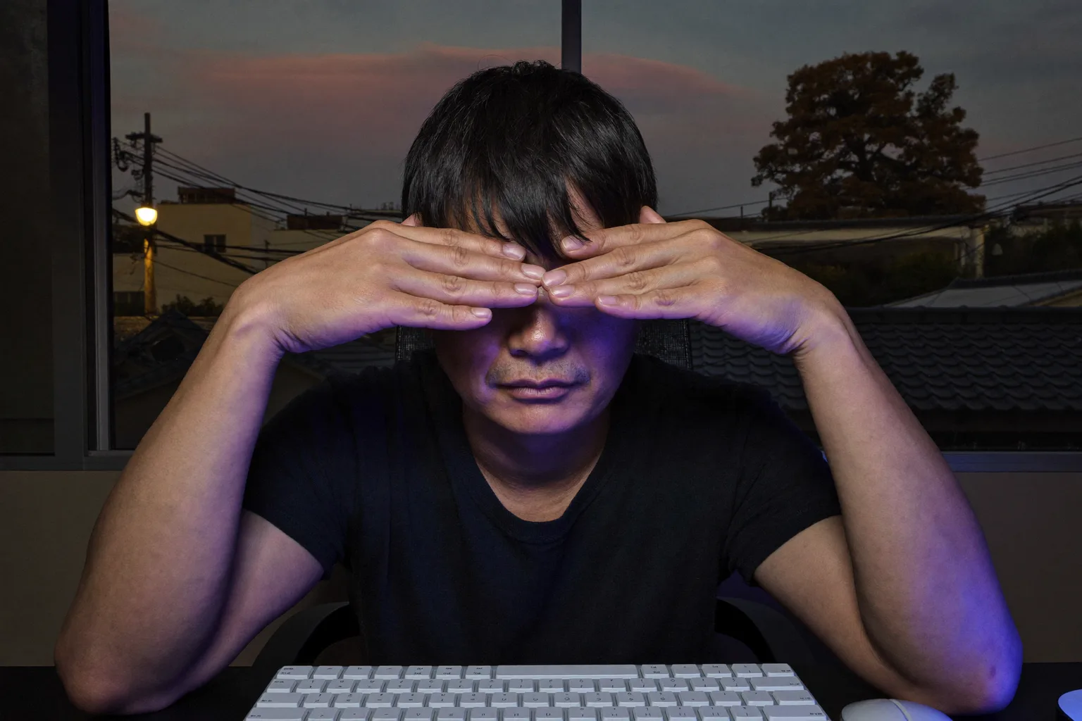 薄暗い部屋のデスクで、閉じた目に手のひらを当てて眼精疲労をやわらげる男性。顔はノートパソコンの画面に照らされている