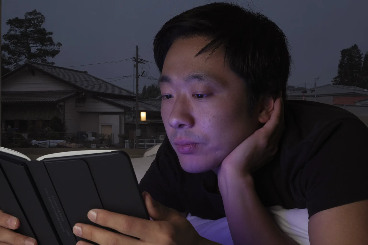 夜遅く、ベッドで電子書籍リーダーを読む男性。画面のかすかな光は顔にほとんど届いていない。かたわらのナイトテーブルでは、画面を上にして置かれたスマートフォンが、隣のランプシェードを冷たく強いブルーライトで照らしている