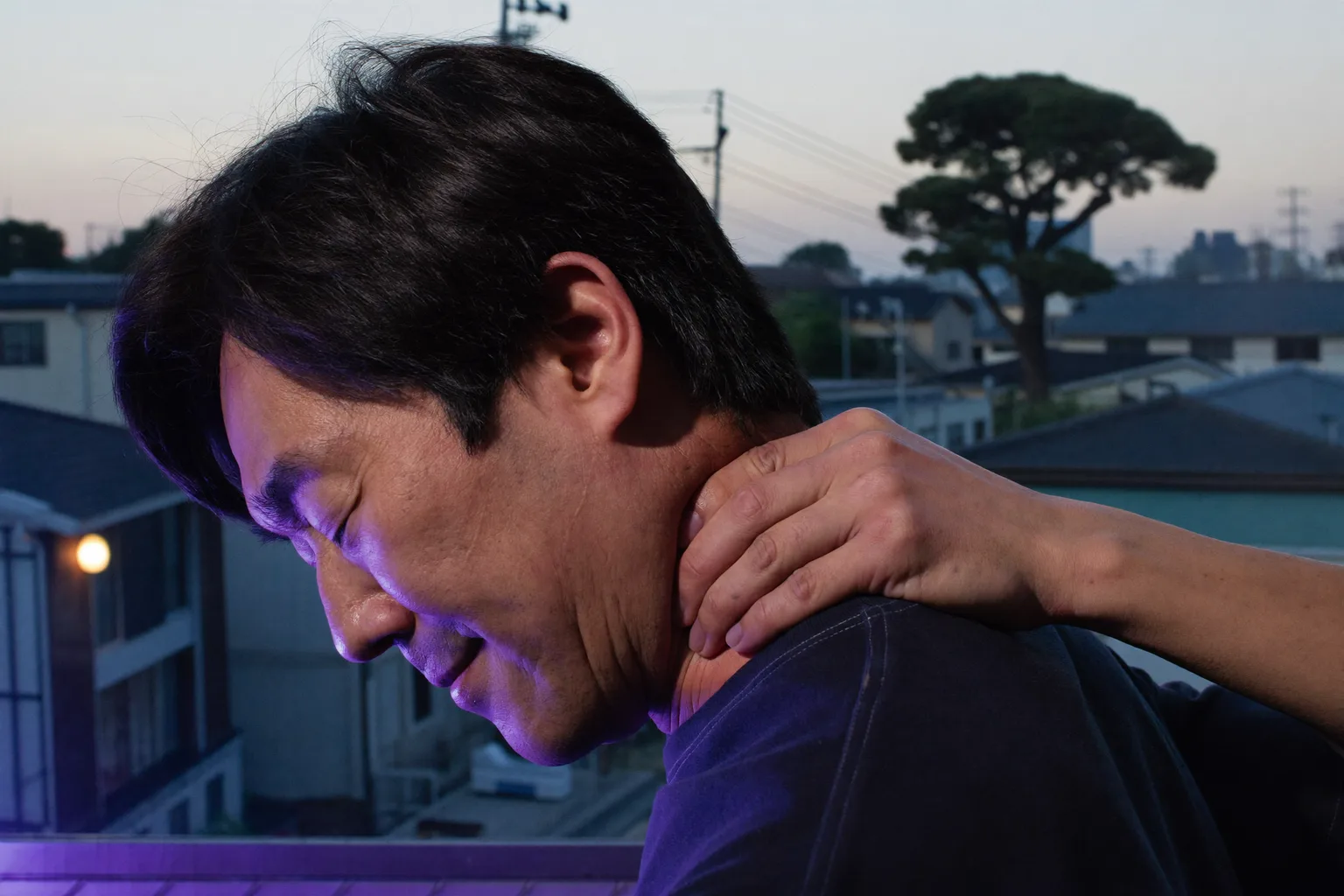 デスクでノートパソコンの前に座り、後ろに手を回して首の付け根をさすっている人。長時間のパソコン作業でこわばり痛む首を表している