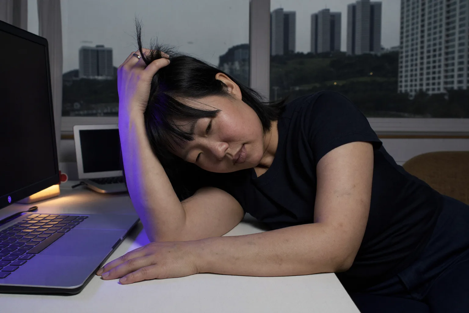 늦은 저녁, 집 책상 앞에 앉은 사람이 한 손으로 머리를 감싸 쥔 채 고개를 기울이고 있다. 시선은 아래로 향한 채 초점이 흐려져 있고, 다른 팔은 텅 빈 책상 위에 가만히 놓여 있다