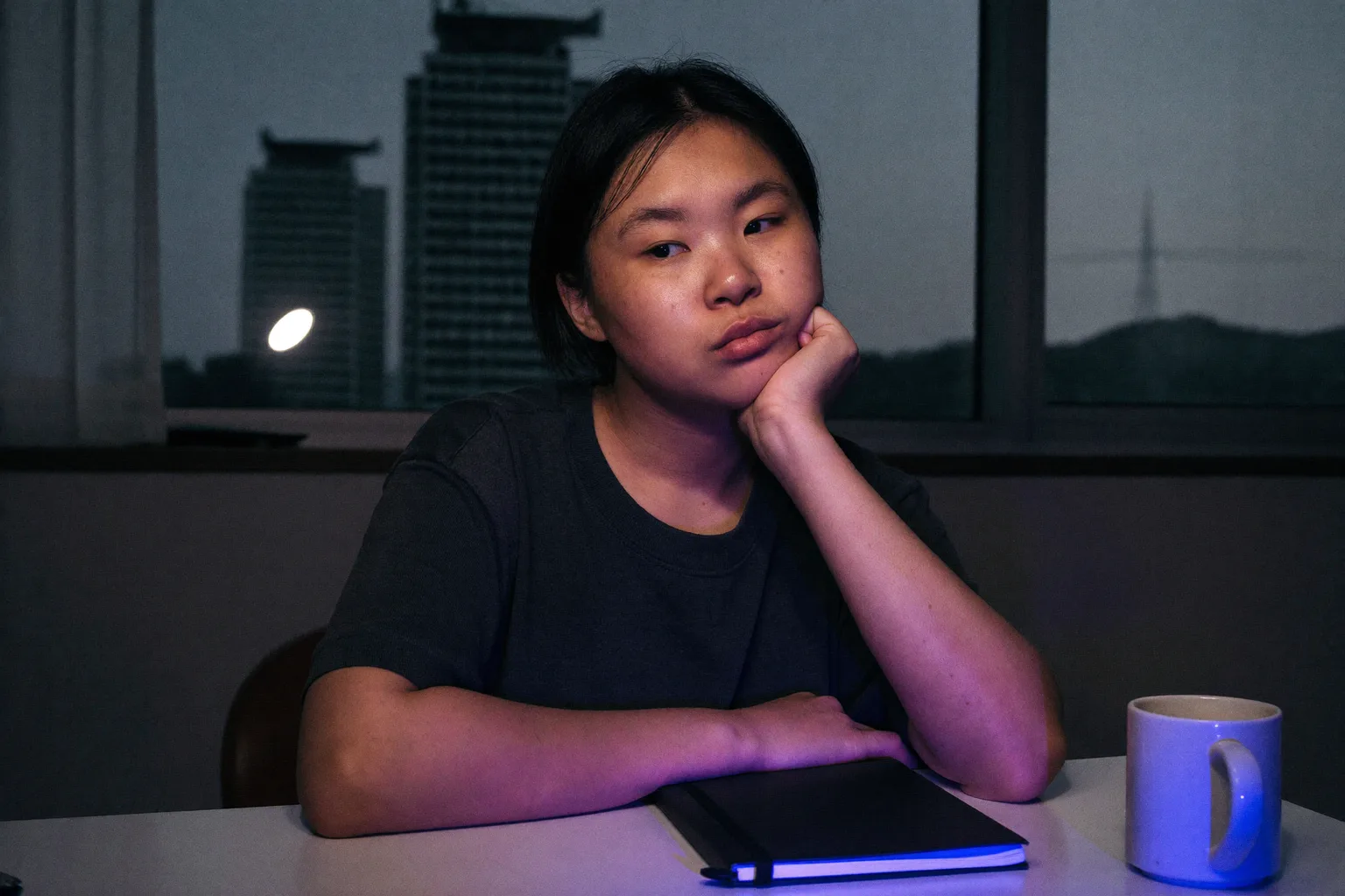 늦은 저녁, 책상 앞에 앉은 학생이 한 손바닥에 턱을 괸 채, 화면 불빛이 옆에서 얼굴을 비추고, 시선은 공부에서 벗어나 프레임 밖으로 흘러가 있다