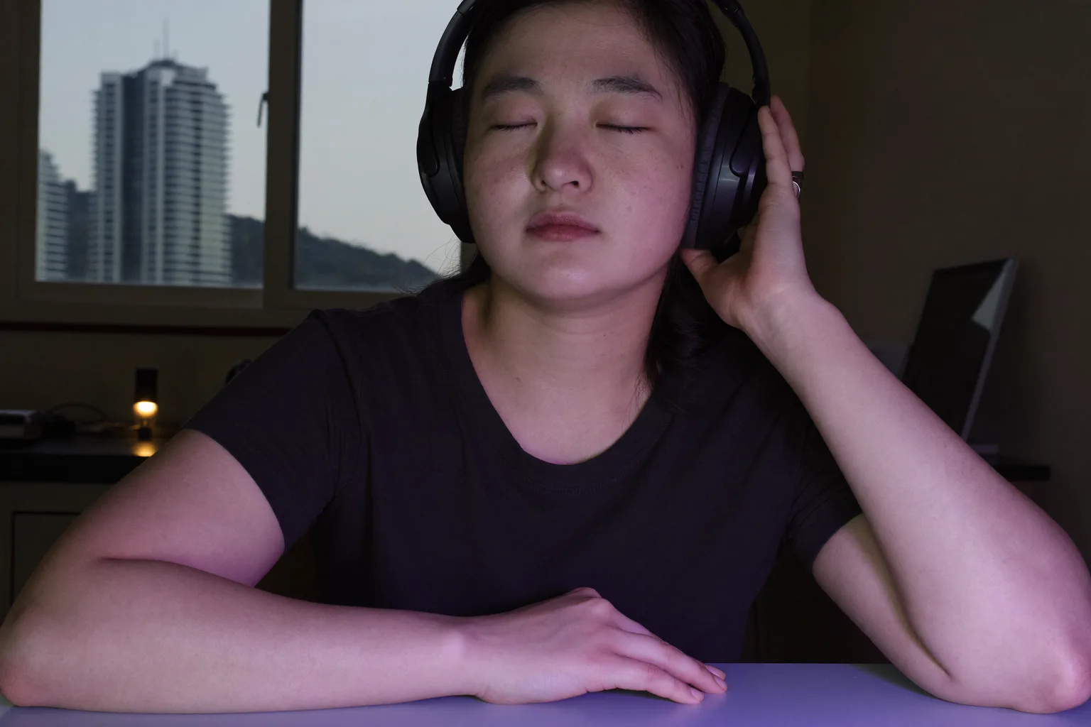 책상 앞에서 오버이어 헤드폰을 쓴 사람이 눈을 감고 한 손을 이어컵에 얹은 채, 음악을 들으며 깊이 몰입해 일하고 있다