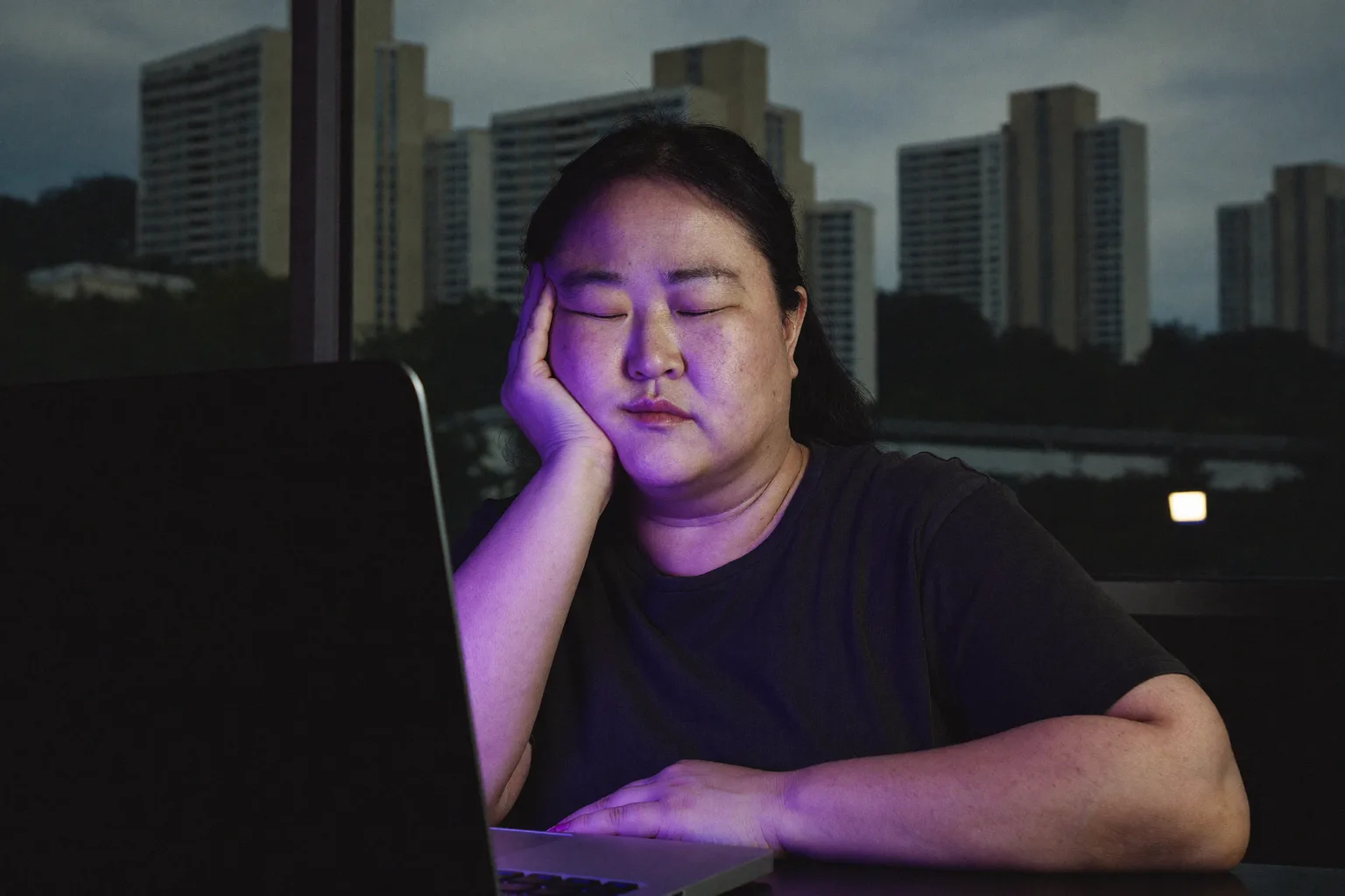 하루가 저물 무렵 노트북 앞에서 손으로 턱을 괸 채 지친 여성이 어둑한 방 안에서 무겁고 건조한 눈과 다크서클을 보이고 있는 모습