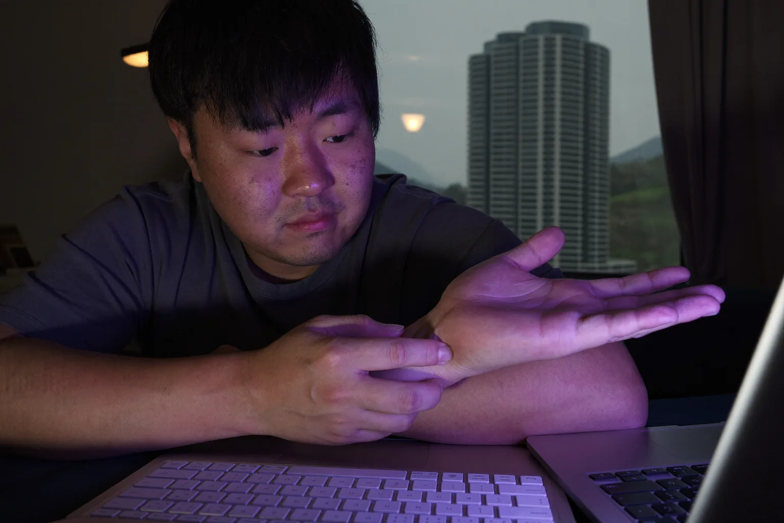 책상에 앉아 다른 손으로 오른쪽 손목을 감싸 쥔 사람, 앞에는 키보드가 있고, 손목터널증후군의 저리고 아픈 손을 보여주는 모습