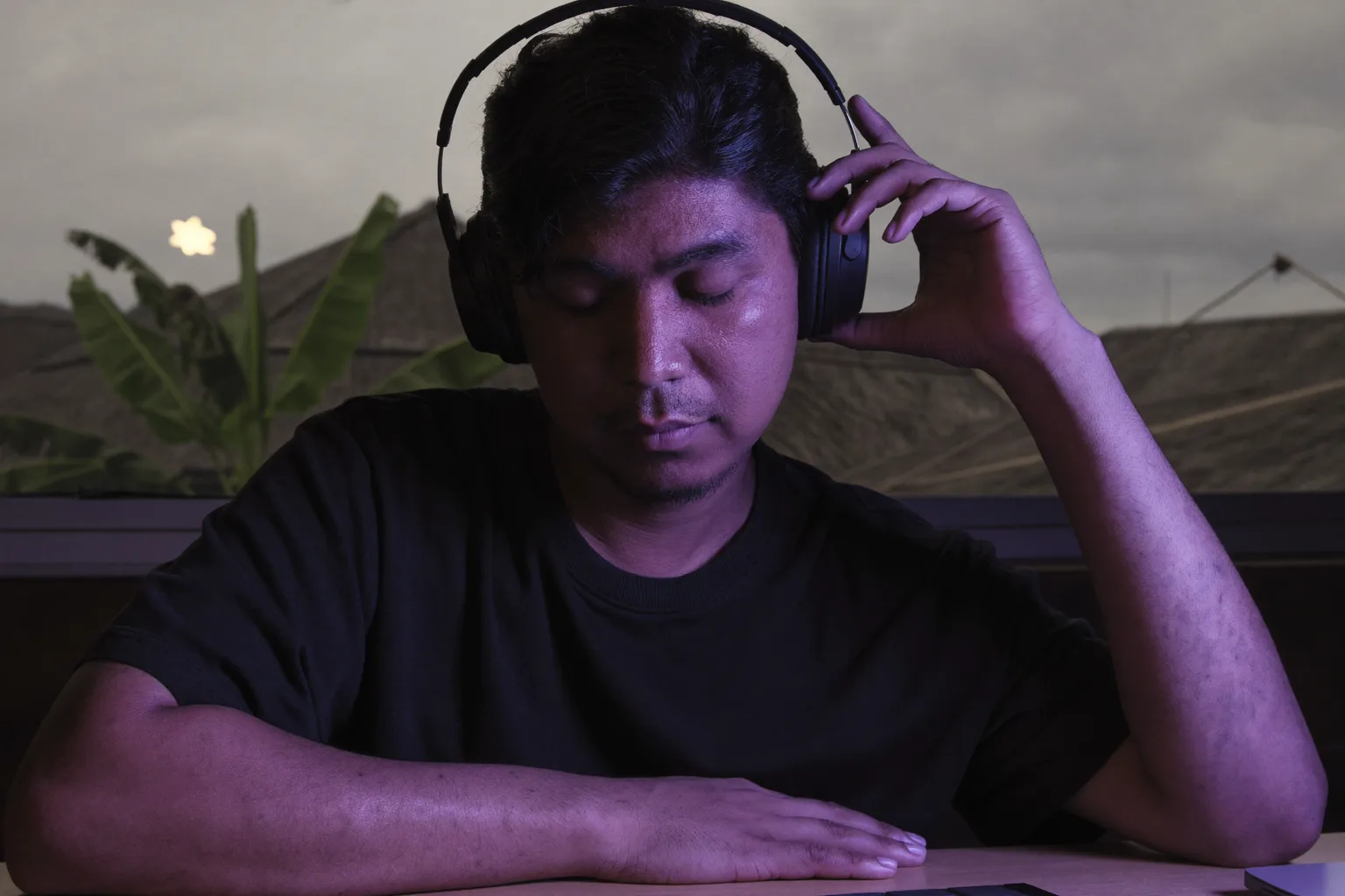 Seseorang di meja kerja memakai fon kepala over-ear, mata tertutup dengan sebelah tangan berehat di atas but fon telinga, sedang khusyuk dalam sesi kerja sambil muzik dimainkan