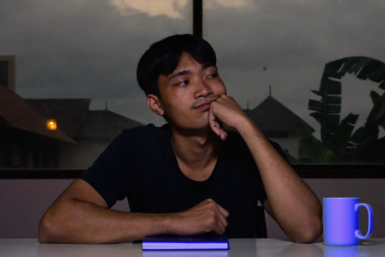 Seorang pelajar di meja pada waktu malam, dagu disandarkan pada satu tapak tangan terbuka, wajah disuluh cahaya skrin dari sisi, pandangan terleret jauh daripada kerja dan keluar dari bingkai