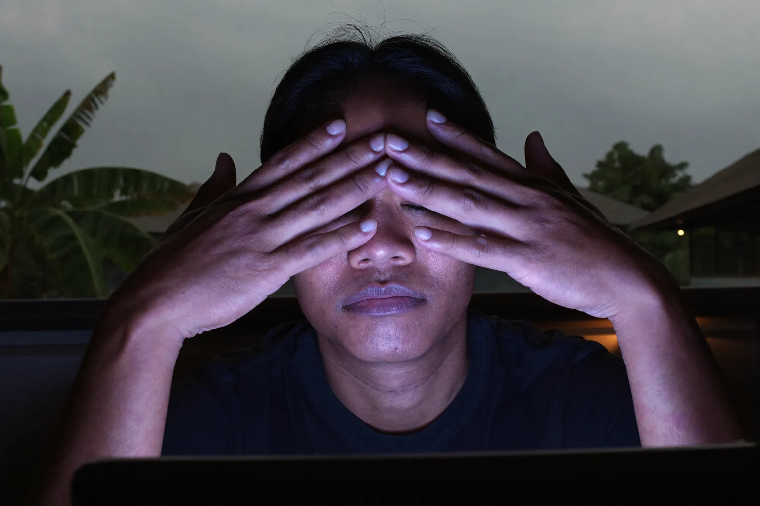 Seorang lelaki di meja kerja dalam bilik malap, menudungkan tapak tangan pada matanya yang tertutup untuk meredakan mata penat, wajahnya disuluh cahaya skrin komputer riba