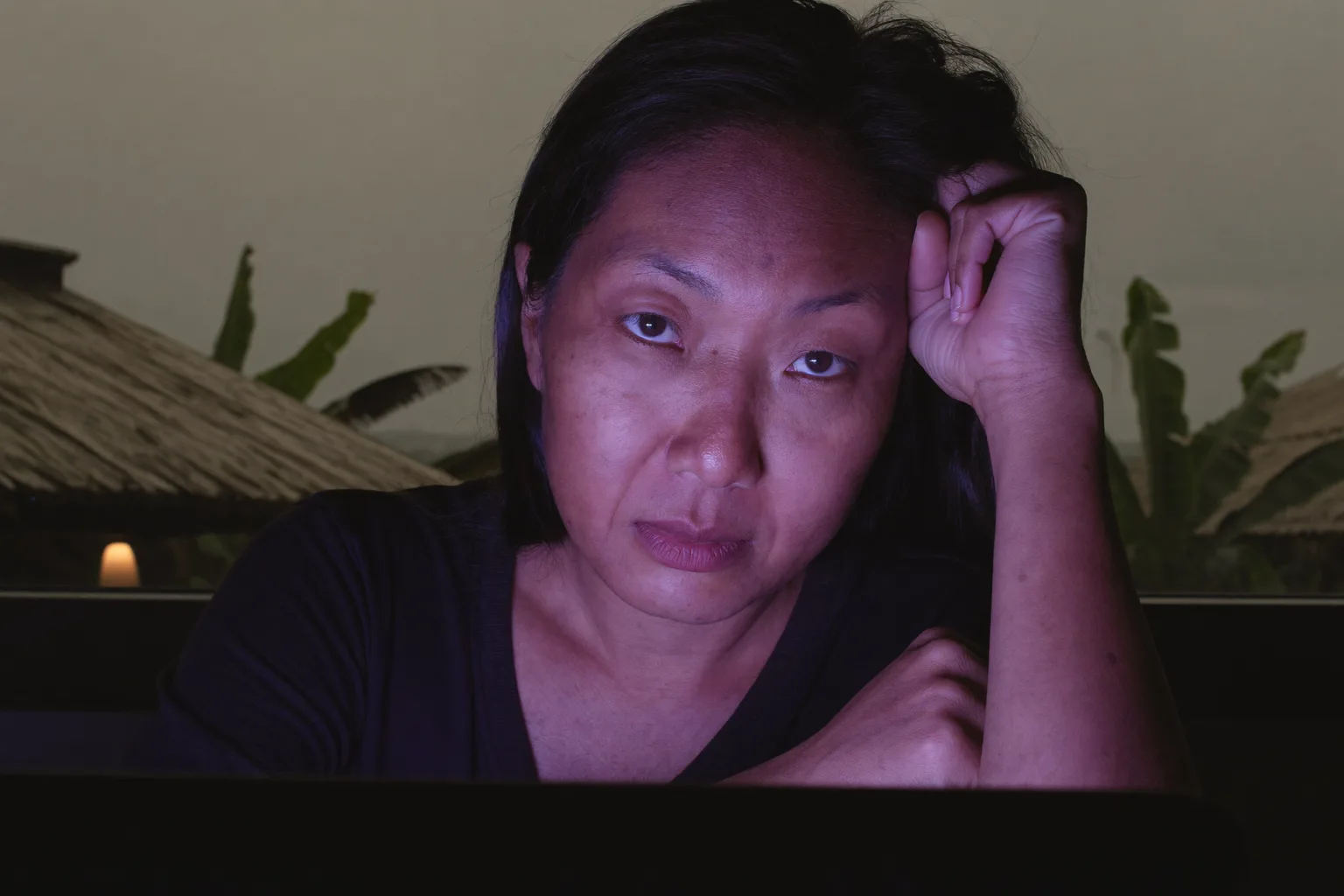 Seorang wanita yang keletihan di hadapan komputer riba lewat hari, mengalas kepala pada tangannya, dengan mata berat, kering dan kelibat gelap di bawah mata dalam bilik yang malap cahayanya