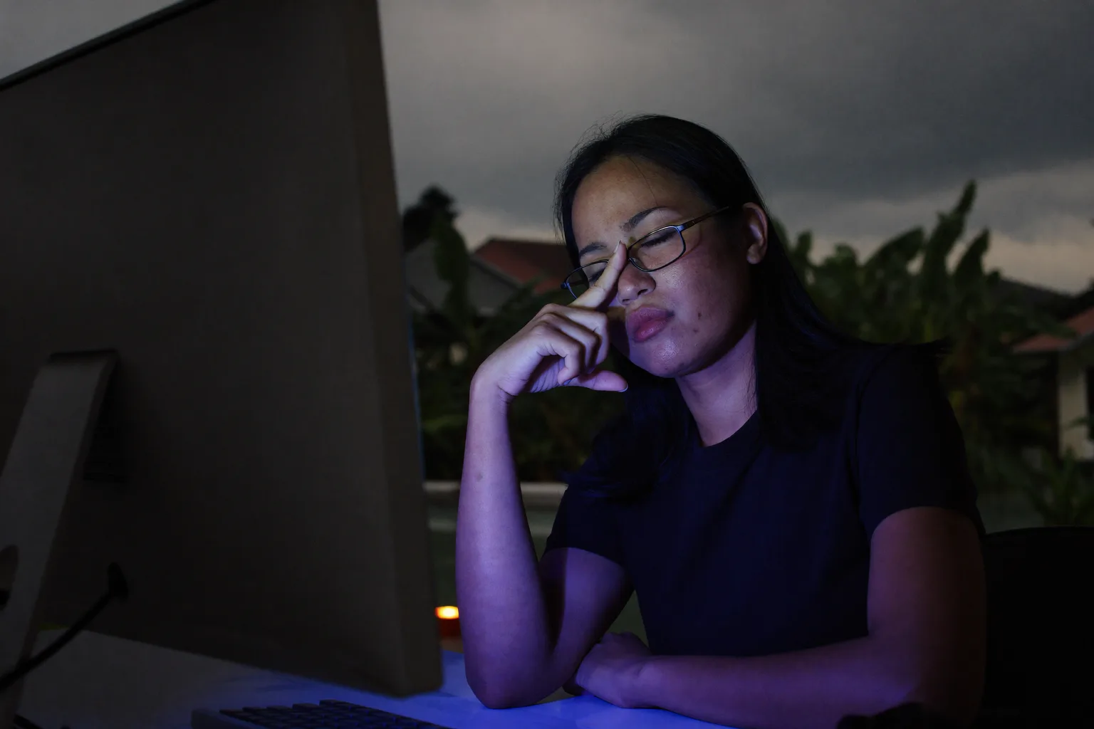 Seorang wanita di meja kerja dalam bilik gelap, mata tertutup dan cermin matanya diselak ke dahi, memicit pangkal hidung di belakang skrin monitornya, wajahnya disuluh cahaya biru sejuk daripada skrin