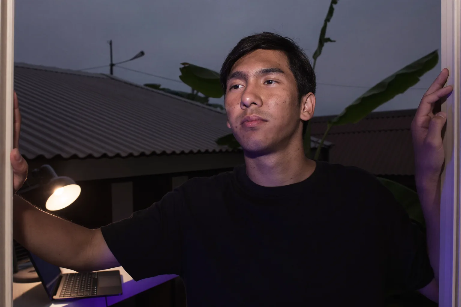 Komputer riba yang ditutup di atas meja kerja di rumah pada waktu malam, dengan lampu bilik menyala dan seseorang berjalan pergi, menggambarkan penghujung hari bekerja dari rumah
