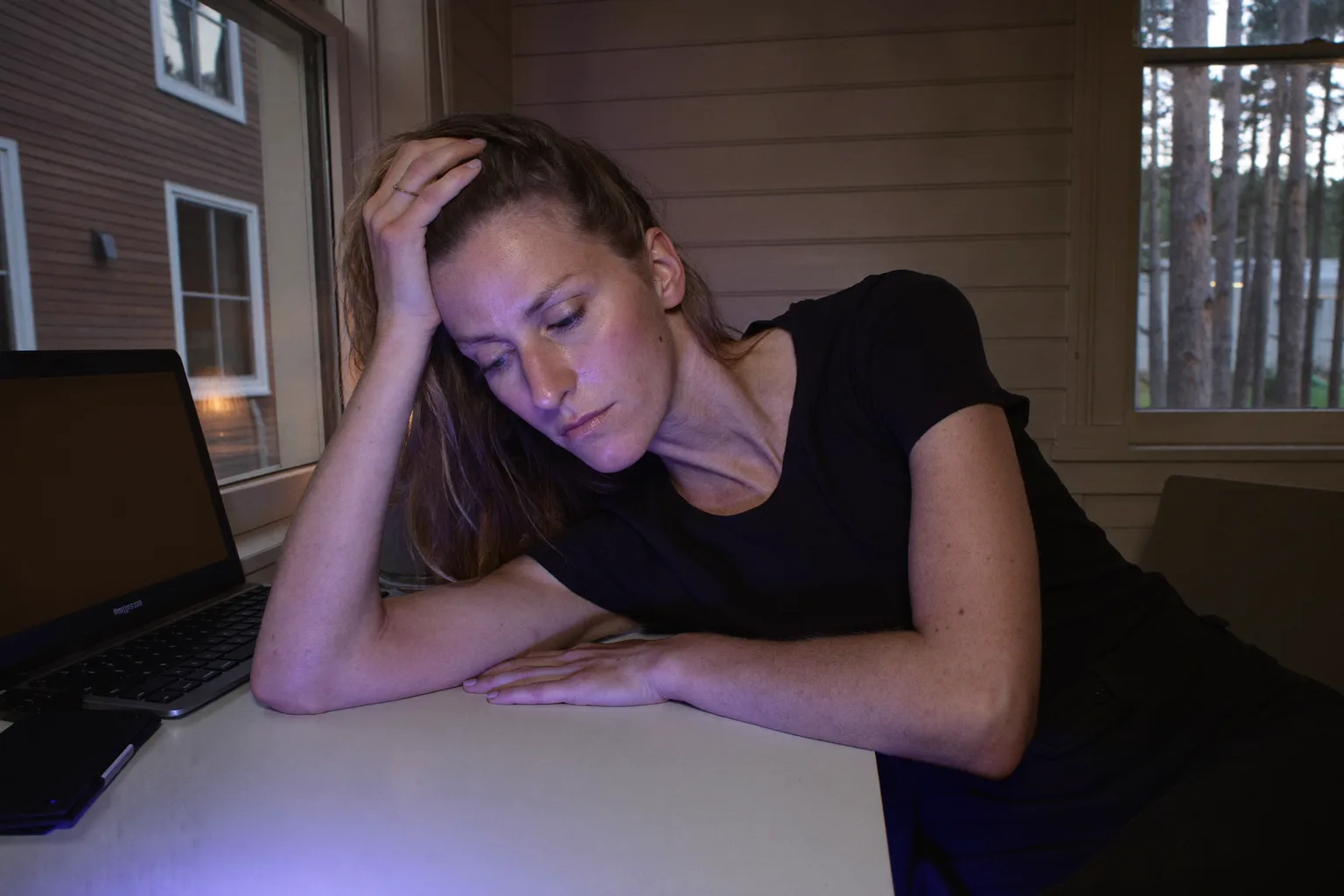 En person ved et hjemmekontor sent på kvelden, med hodet lent inn i den ene hånden og fingrene i håret, blikket senket og ufokusert, mens den andre underarmen ligger stille på et tomt skrivebord