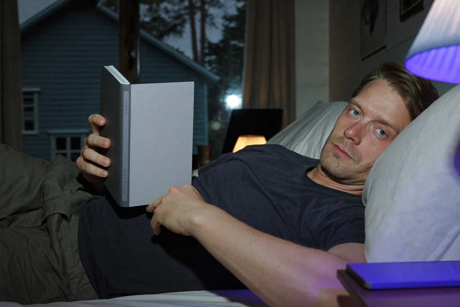 En mann som leser på en e-bokleser i sengen sent på kvelden, siden lyser svakt og når så vidt ansiktet hans, mens en mobil som ligger med skjermen opp på nattbordet ved siden av, fyller den andre lampeskjermen med hardt, kaldt blått lys