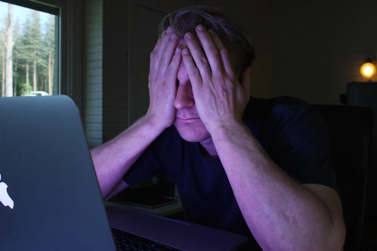 En mann ved et skrivebord i et dunkelt rom presser håndflatene mot de lukkede øynene for å lindre øyebelastning, ansiktet opplyst av laptopskjermen