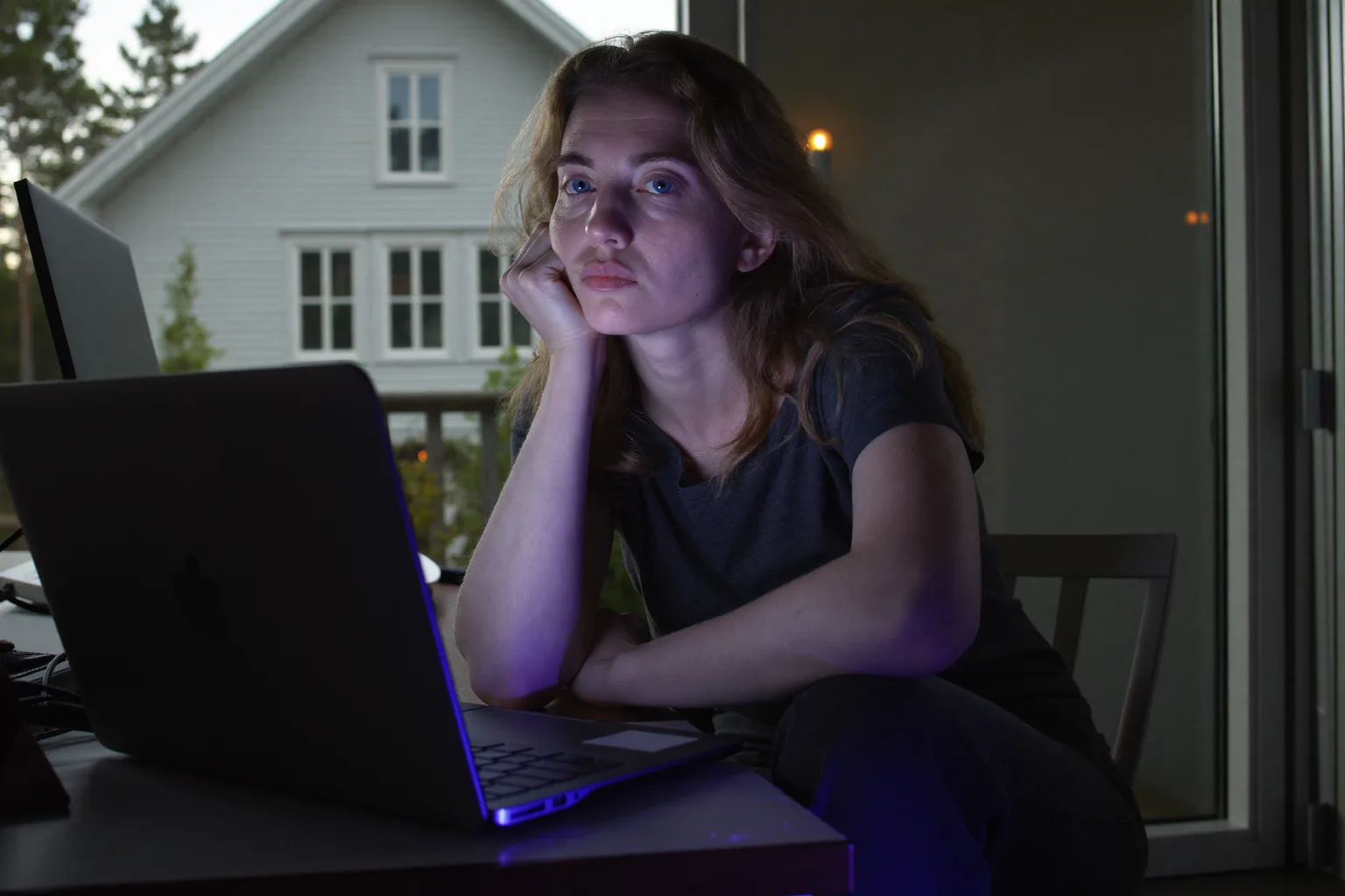 En sliten kvinne ved en bærbar PC sent på dagen, som hviler hodet i hånden, med tunge, tørre øyne og mørke ringer i et svakt opplyst rom