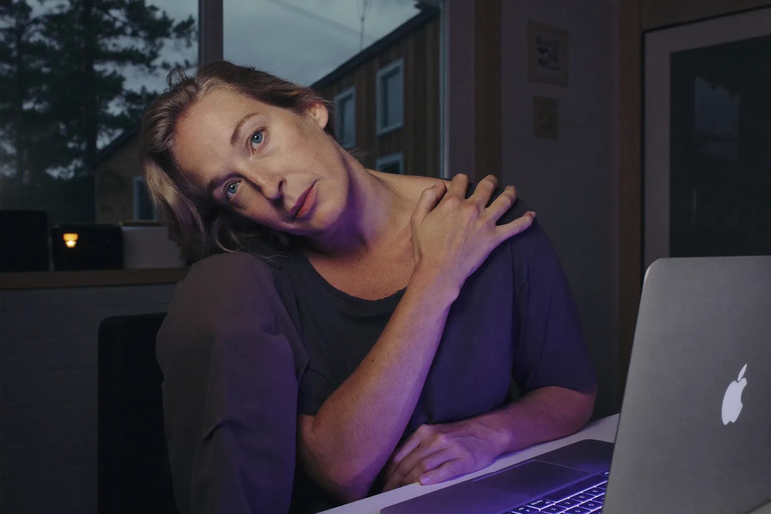 En person ved et skrivebord som strekker seg bakover for å gni nedre del av nakken foran en bærbar PC, som viser den stive, verkende nakken etter en lang dag foran skjermen