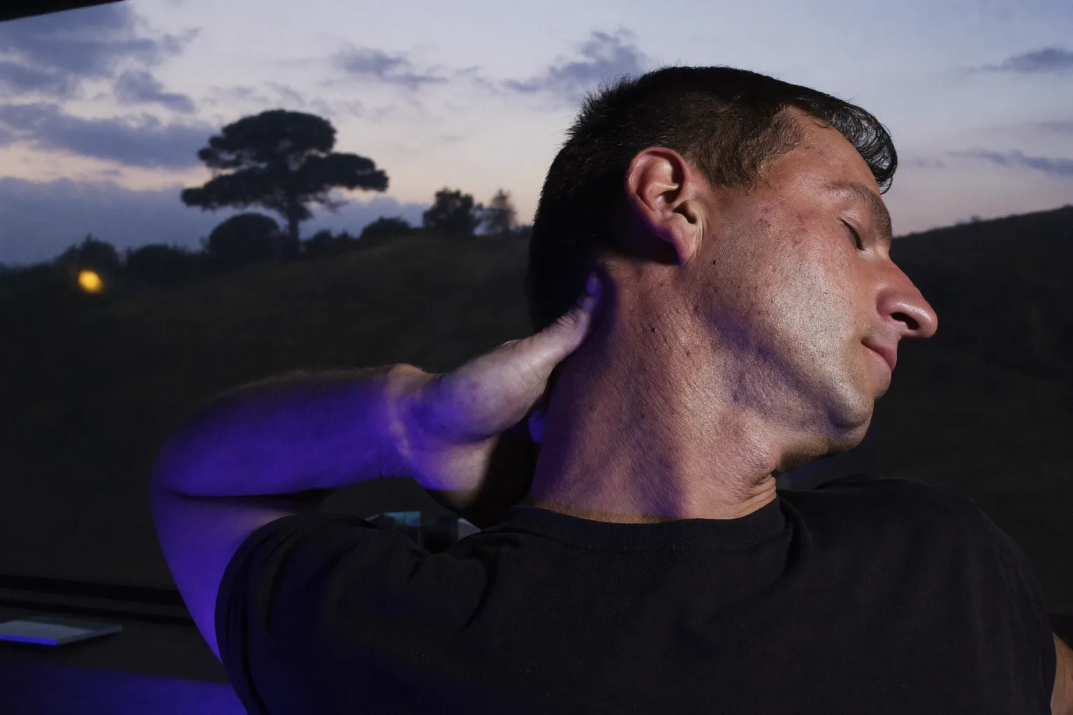 A person at a desk reaching back to rub the base of their neck in front of a laptop, showing the stiff, aching neck of a long computer day