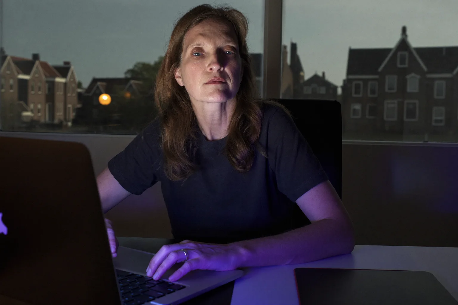 Iemand die laat op de avond aan een bureau thuis zit, met de blik voorbij het laptopscherm en de handen nog op het toetsenbord, de vlakke uitputting van een burn-out op het werk