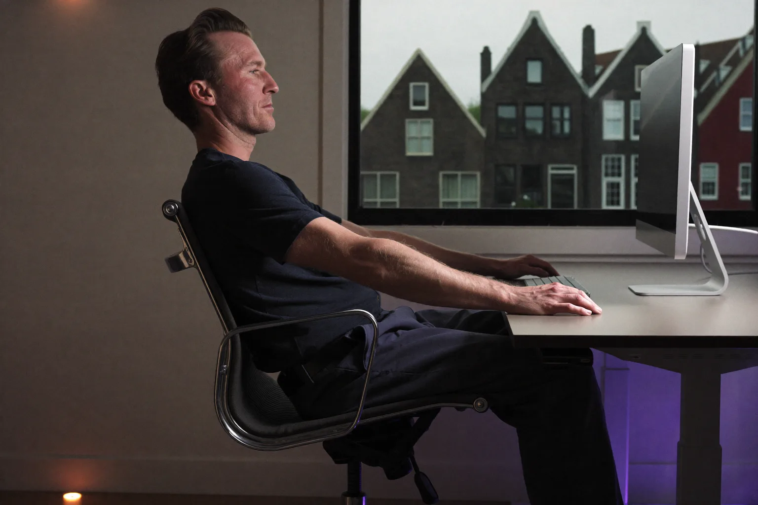Een persoon van opzij gezien, zittend aan een bureau met de voeten plat op de vloer, de rug tegen de rugsteun van de stoel, de onderarmen op bureauhoogte en het beeldscherm op ooghoogte: de referentiehouding achter een bureau