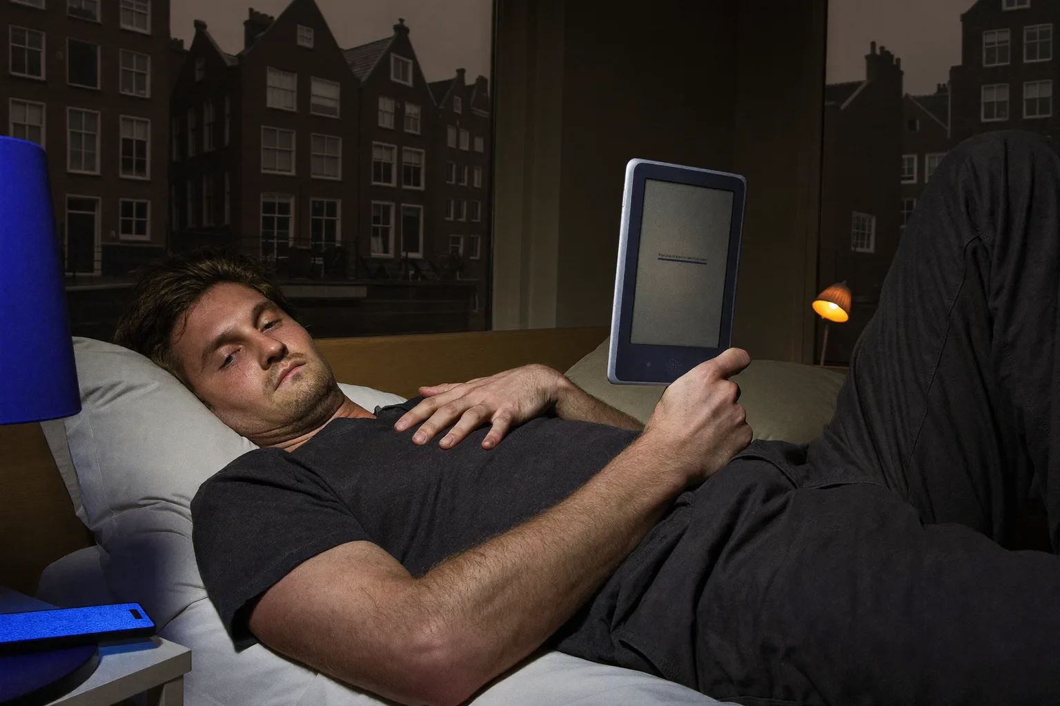Een man leest laat op de avond in bed op een e-reader; de pagina geeft een zwakke gloed af die zijn gezicht amper bereikt, terwijl op het nachtkastje naast hem een telefoon met het scherm naar boven de tweede lampenkap overspoelt met hard, koud blauw licht