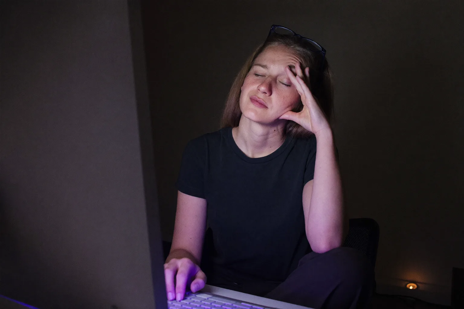 Een vrouw aan een bureau in een donkere kamer, ogen dicht en haar bril omhooggeschoven op haar voorhoofd, terwijl ze met haar vingers haar neusbrug dichtknijpt boven de achterkant van haar monitor, haar gezicht koudblauw verlicht door het scherm