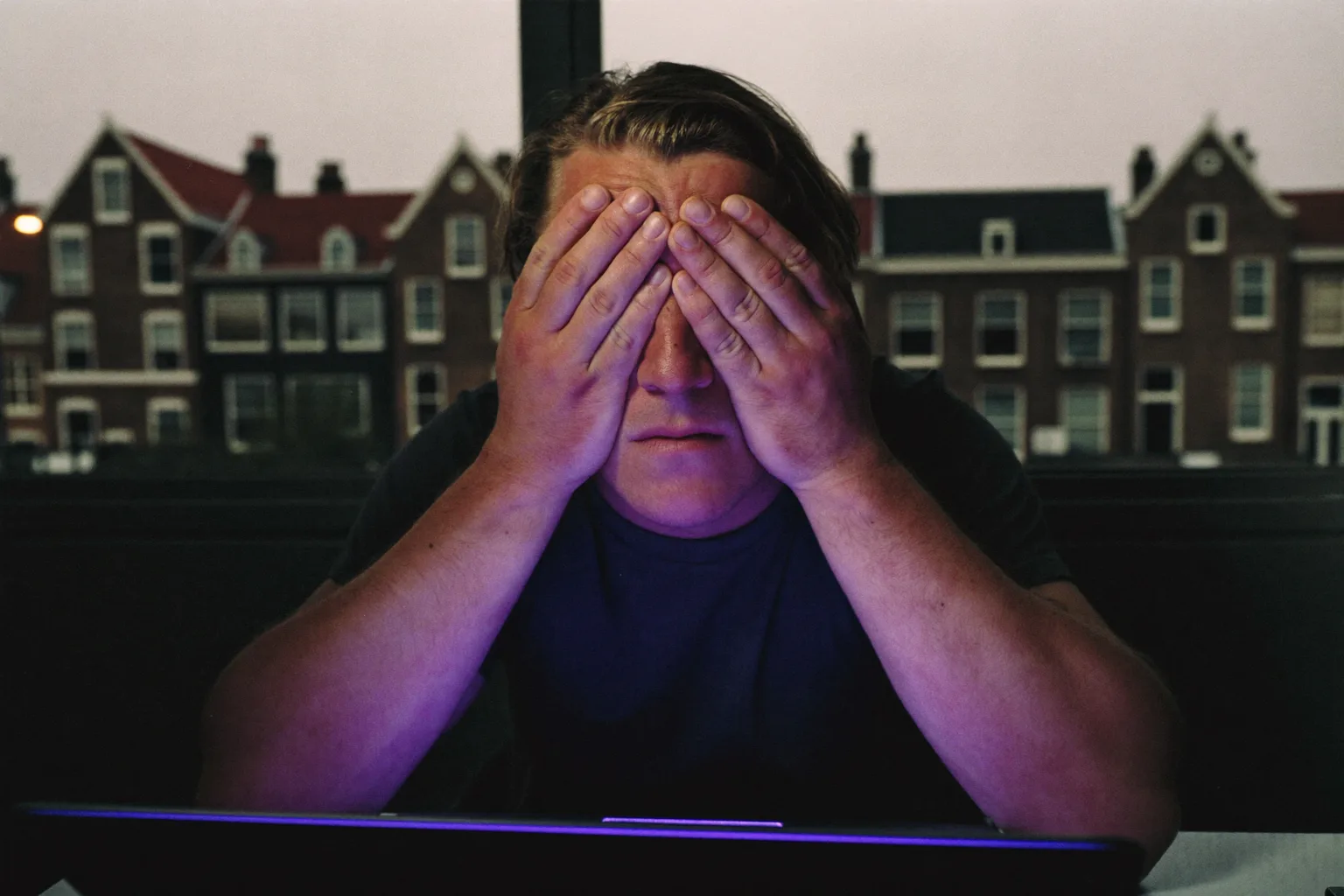 Een man aan een bureau in een schemerige kamer die zijn handpalmen op zijn gesloten ogen drukt om zijn vermoeide ogen te verlichten, zijn gezicht verlicht door het laptopscherm