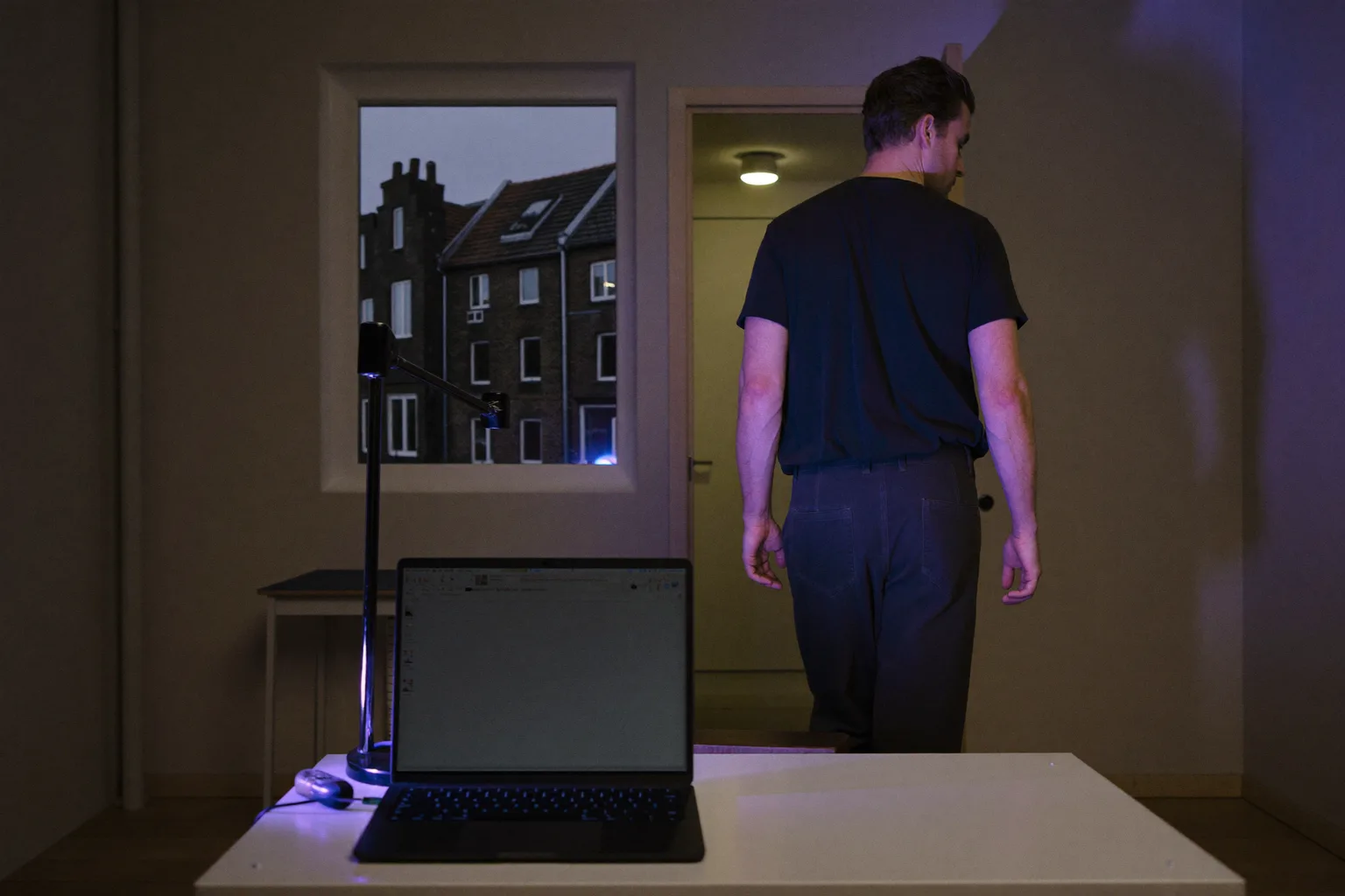 Een dichtgeklapte laptop op een bureau thuis in de avond, met het licht nog aan en iemand die wegloopt: het beeld van het einde van een thuiswerkdag
