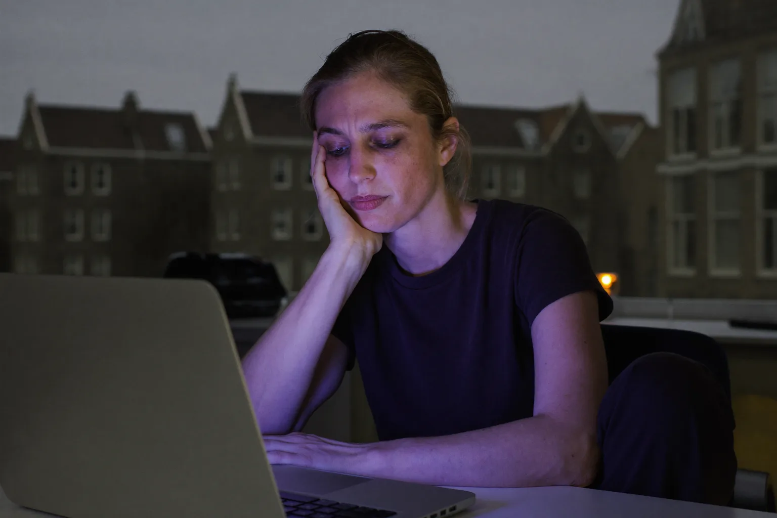Een vermoeide vrouw achter een laptop laat op de dag, met haar hoofd steunend op haar hand, zware, droge ogen en wallen, in een schemerig verlichte kamer