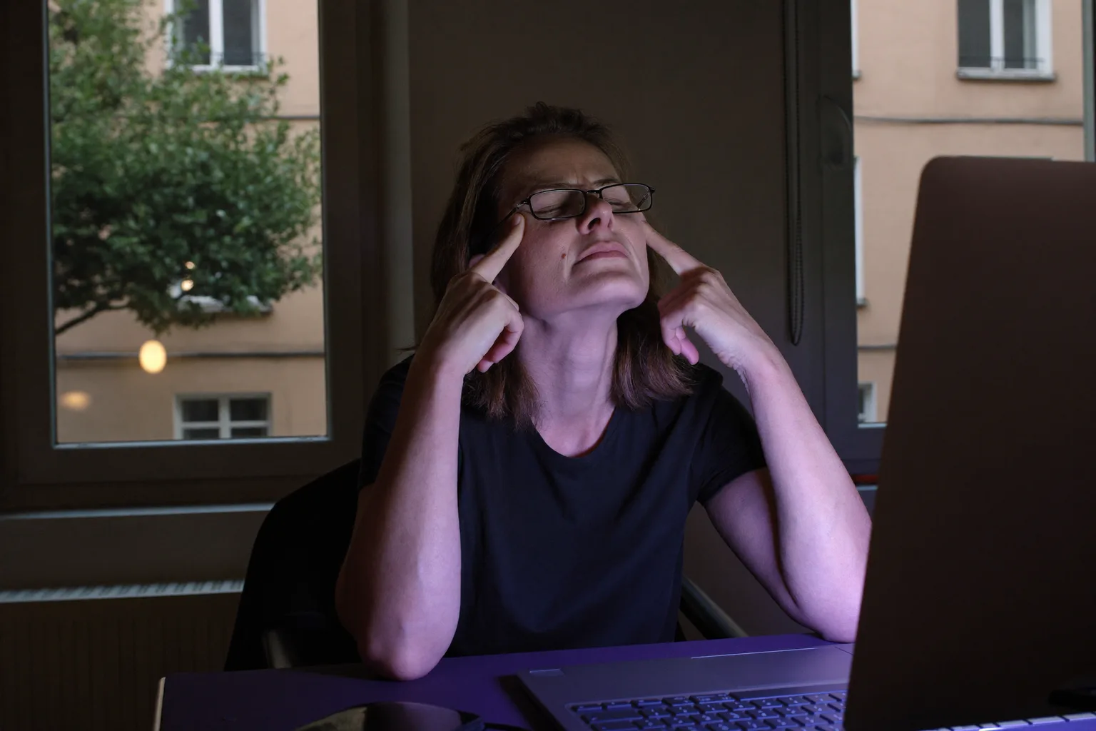 Kobieta przy biurku w ciemnym pokoju, z zamkniętymi oczami i okularami przesuniętymi na czoło, uciskająca nasadę nosa nad tyłem monitora, jej twarz oświetlona zimnym niebieskim blaskiem ekranu