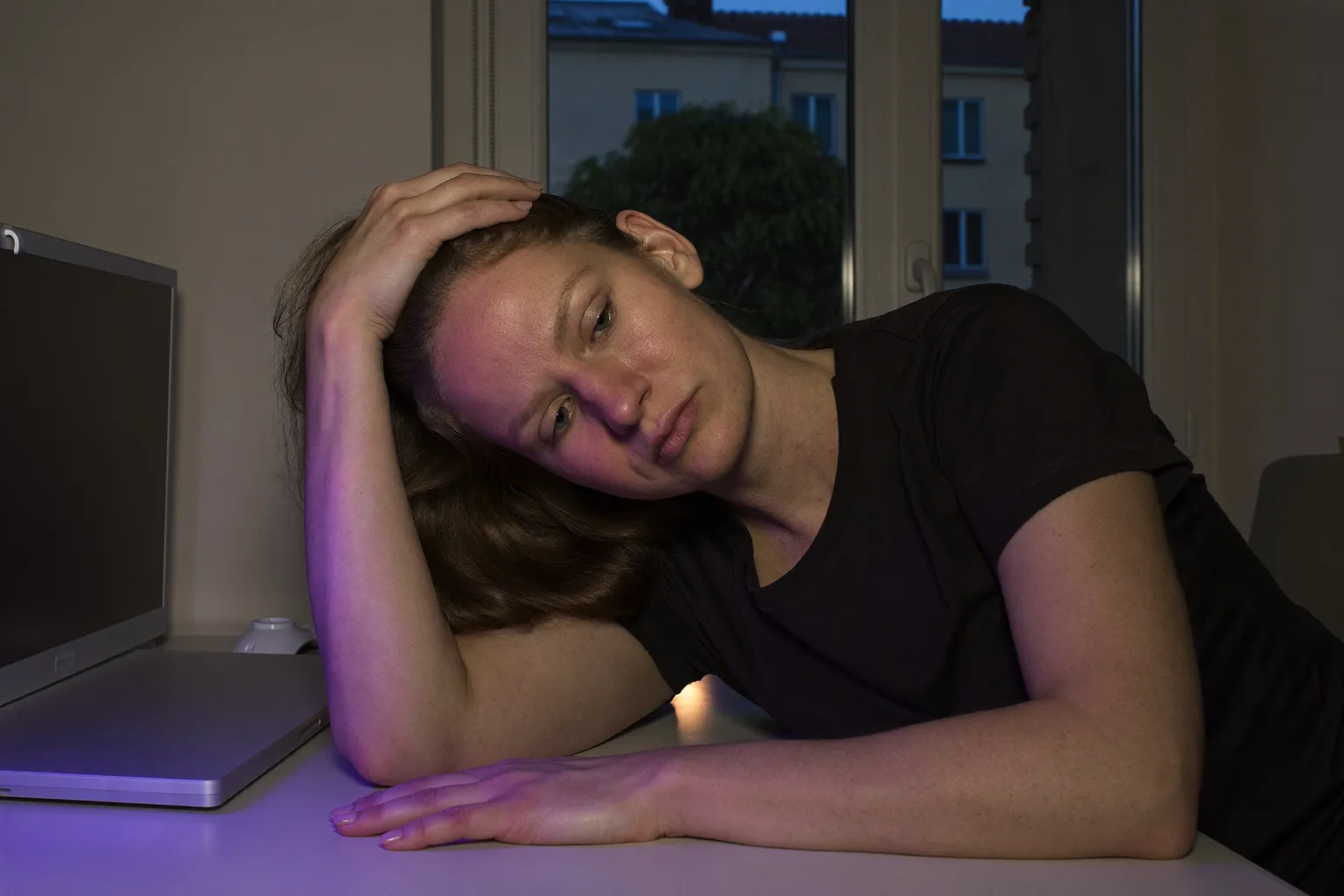 Osoba przy domowym biurku późnym wieczorem, z głową opartą na jednej dłoni, z palcami wplecionymi we włosy, ze wzrokiem spuszczonym i nieostrym, a drugie przedramię leży nieruchomo na pustym blacie