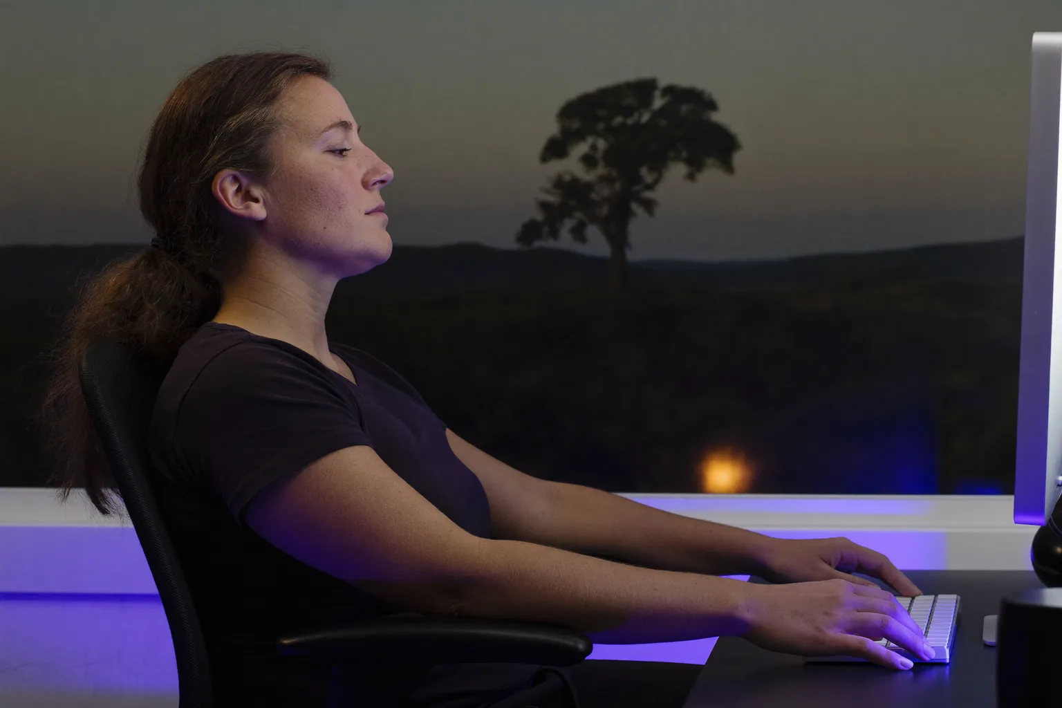 A person seen from the side sitting at a desk with feet flat on the floor, back against the chair support, forearms level with the desk and the monitor at eye level, the reference desk posture