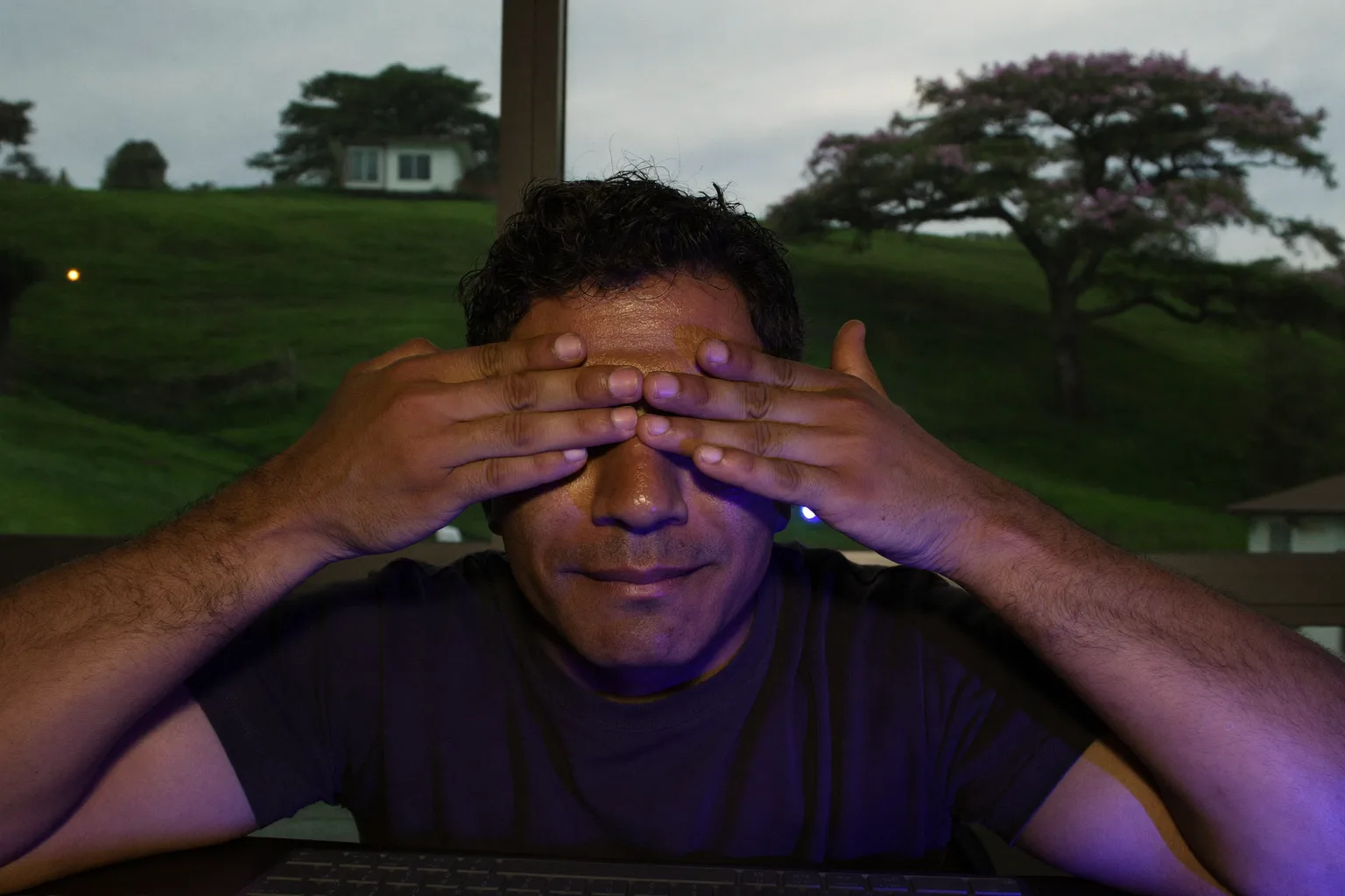 Um homem sentado à mesa, numa sala escura, pressionando as palmas das mãos sobre os olhos fechados para aliviar o cansaço visual e os olhos cansados, o rosto iluminado pela tela do notebook