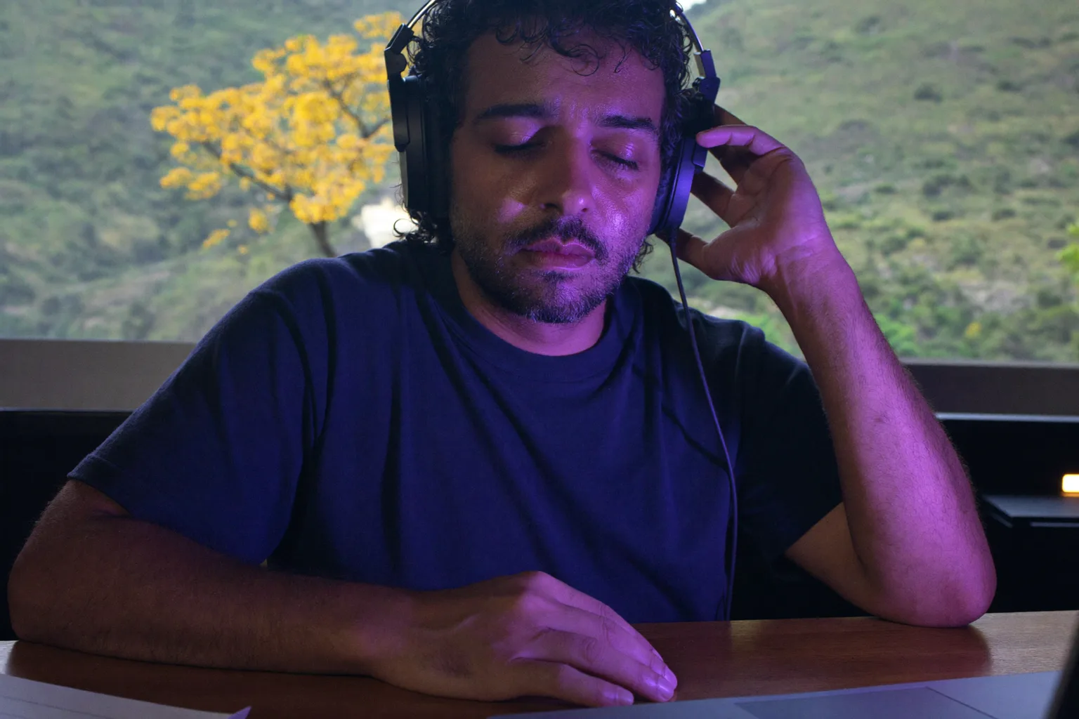 Uma pessoa em uma mesa de trabalho usando fones de ouvido over-ear, olhos fechados e uma mão apoiada na concha do fone, imersa em uma sessão de trabalho com música tocando